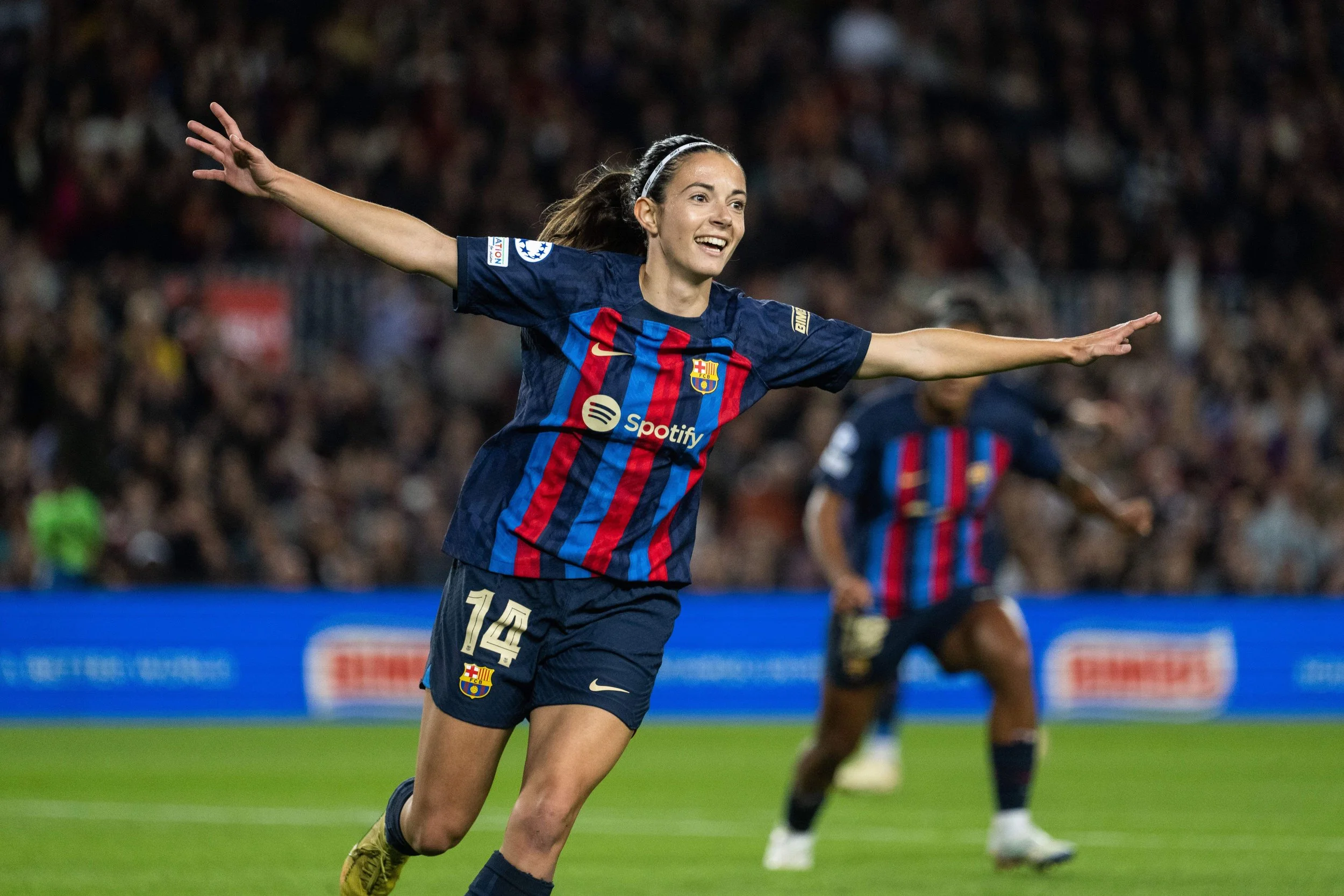 A female soccer player wearing a FC Barcelona jersey celebrating on the field with arms outstretched amidst a crowd of spectators.