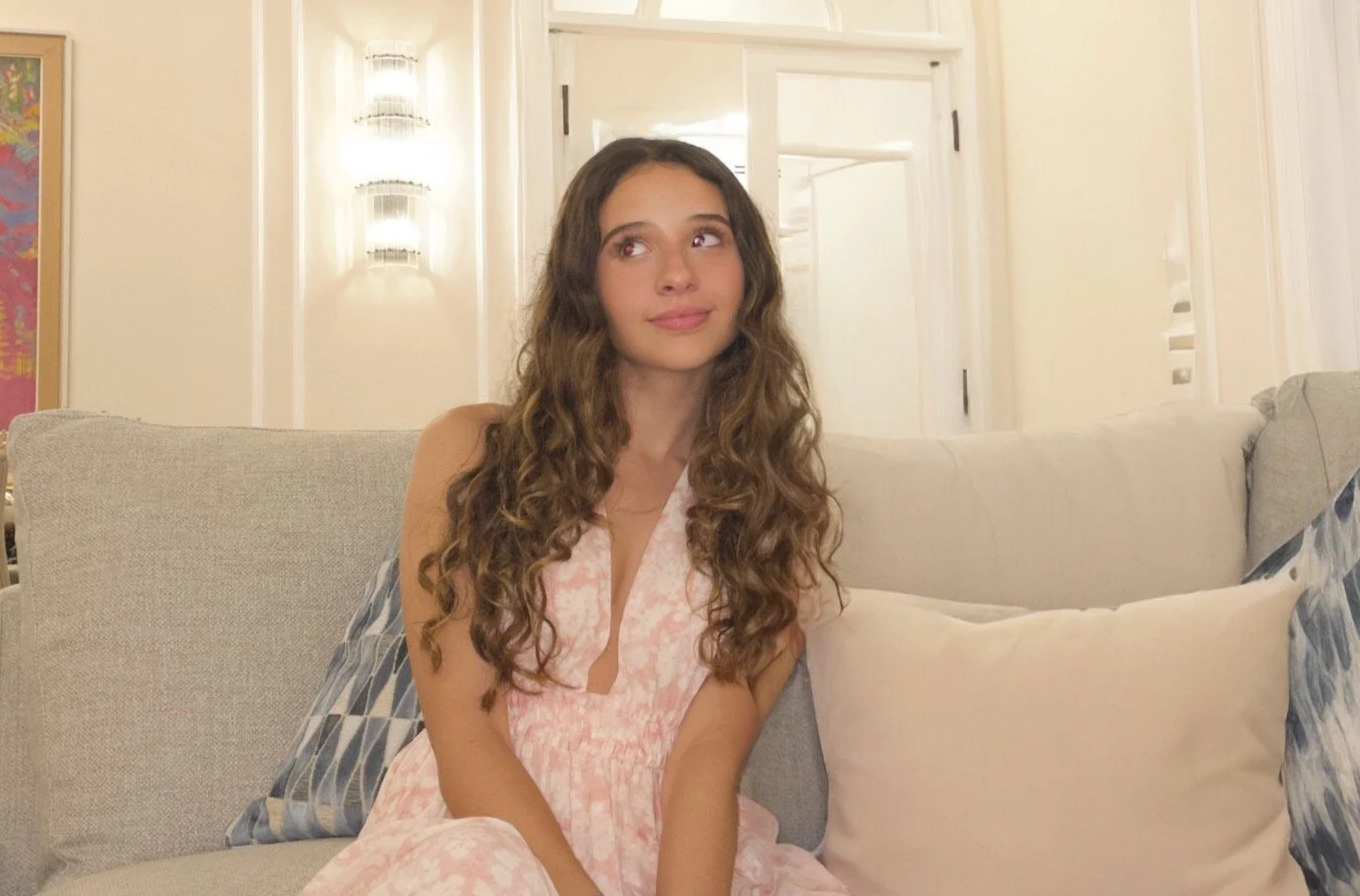 A young woman with long curly brown hair sitting on a beige sofa in a well-lit room, wearing a pink and white sleeveless dress with a keyhole neckline.