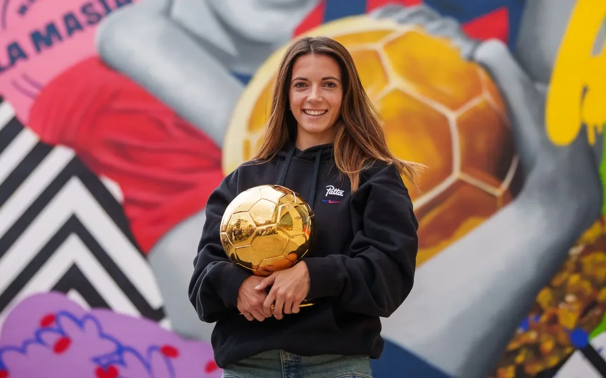 A young woman with long brown hair smiling and holding a gold soccer ball trophy standing outdoors in front of a colorful, graffiti-style mural.