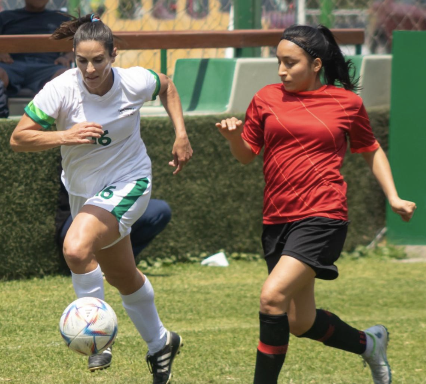 Two female soccer players actively compete for the ball during a match on a grassy field, with a bench and foliage in the background.