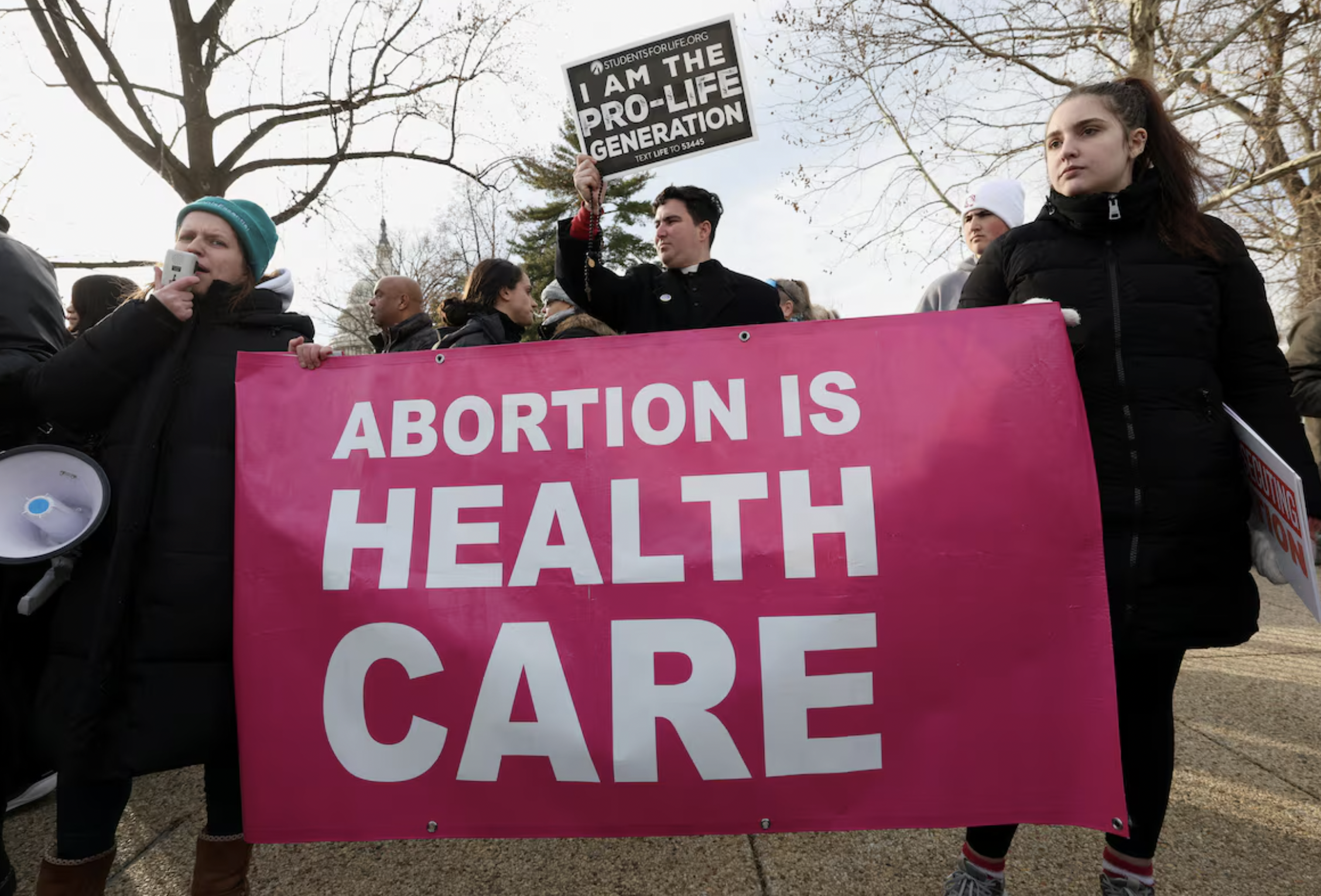 Group of people at a rally holding a large pink banner that reads "ABORTION IS HEAL TH CARE." One person is holding a black and white sign that states "I AM THE PRO-LIFE GENERATION" and mentions a text number for more information. The people are dressed warmly, and leafless trees are visible in the background.