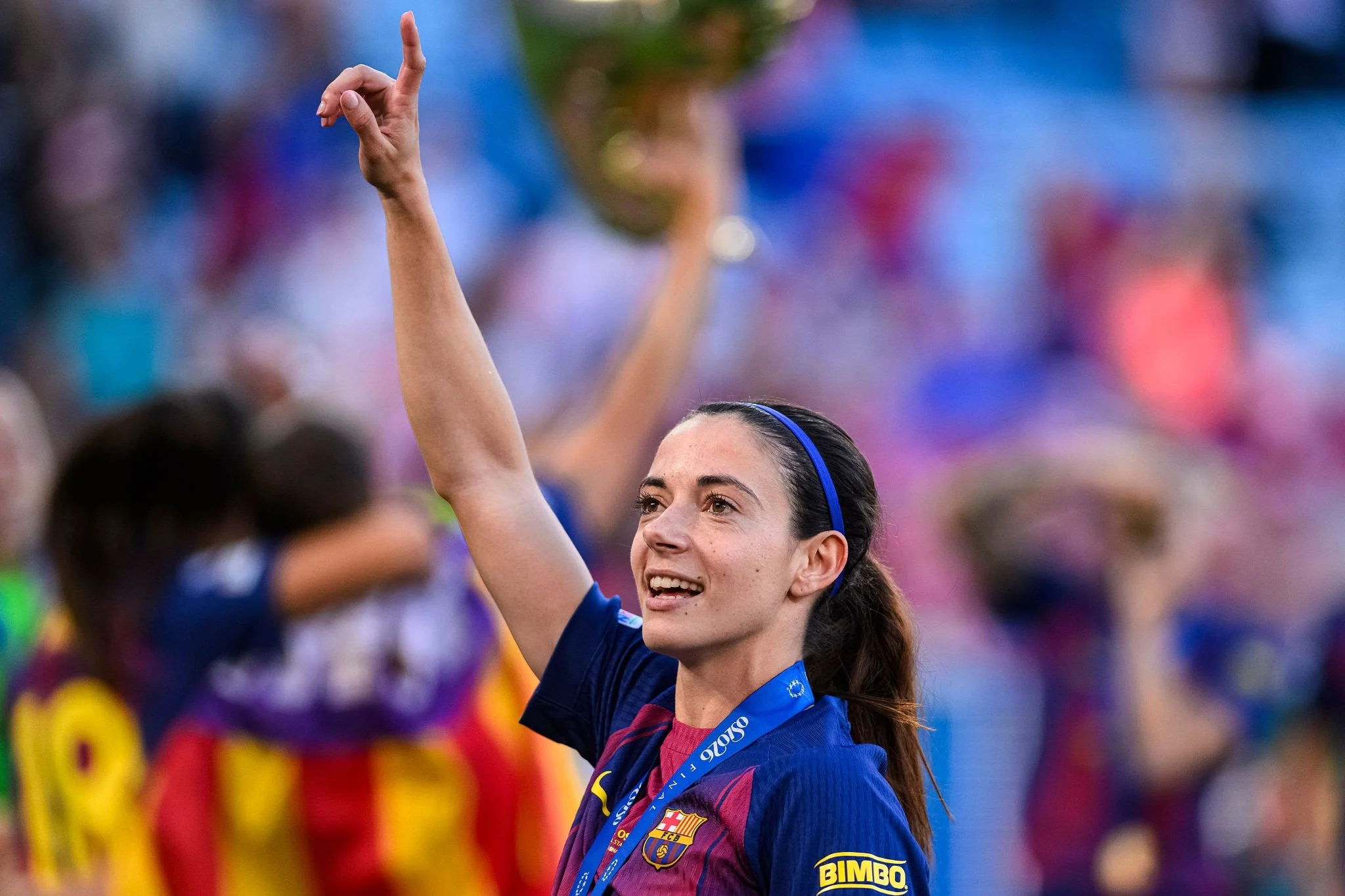 A smiling female soccer player celebrating, wearing a blue and maroon FC Barcelona jersey with Bimbo sponsorship, raising her right hand with index finger pointing up, with colorful blurred crowd in the background.