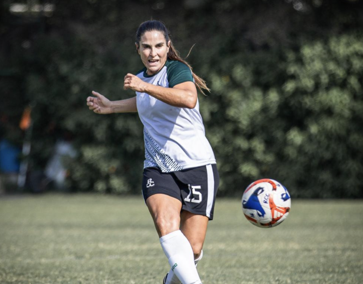 Women soccer player in a white and green jersey running on the field with a soccer ball nearby.