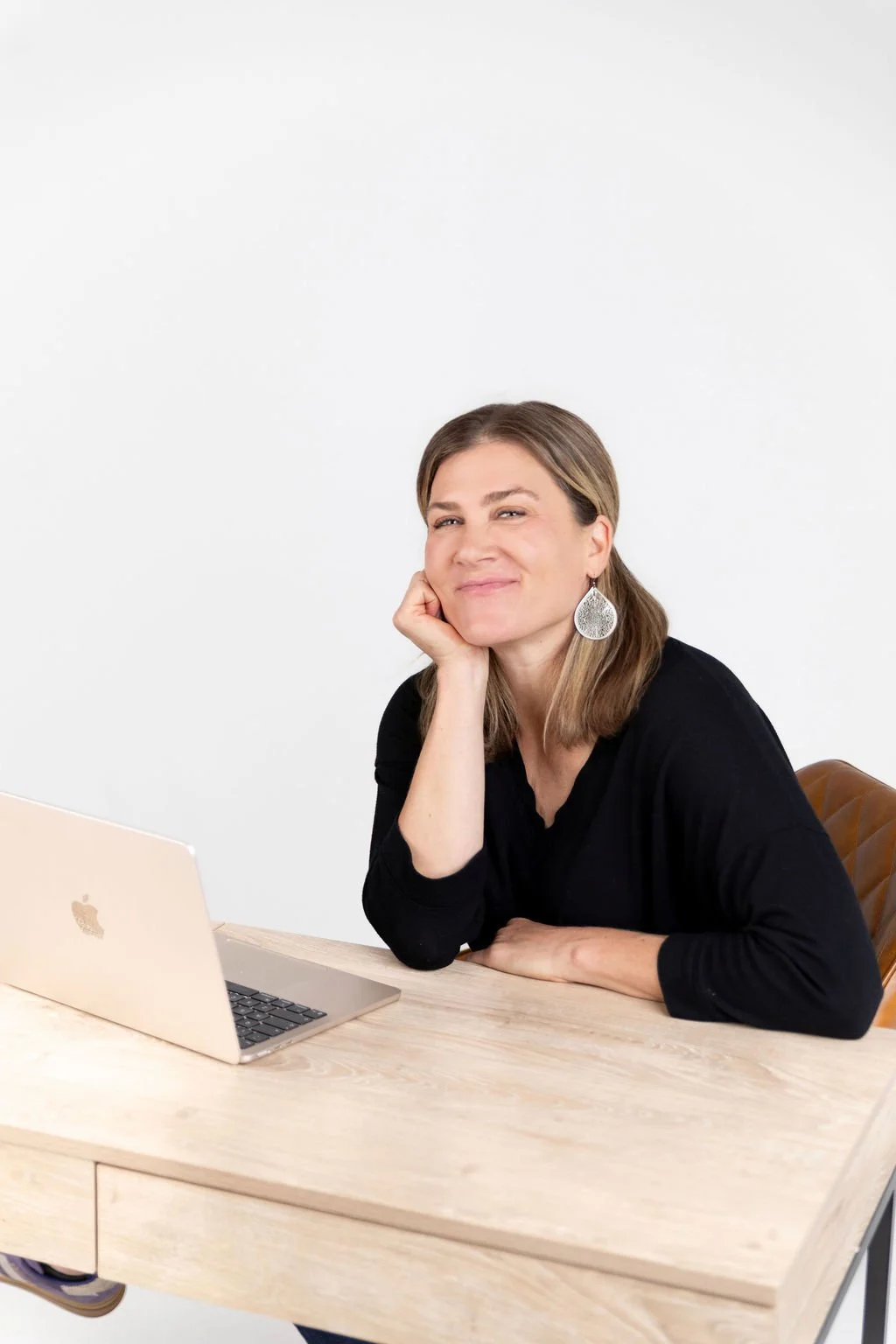 A woman sitting at a light wooden desk with a silver laptop, resting her chin on her hand, smiling at the camera, wearing a black top and large silver earrings, against a plain white background.