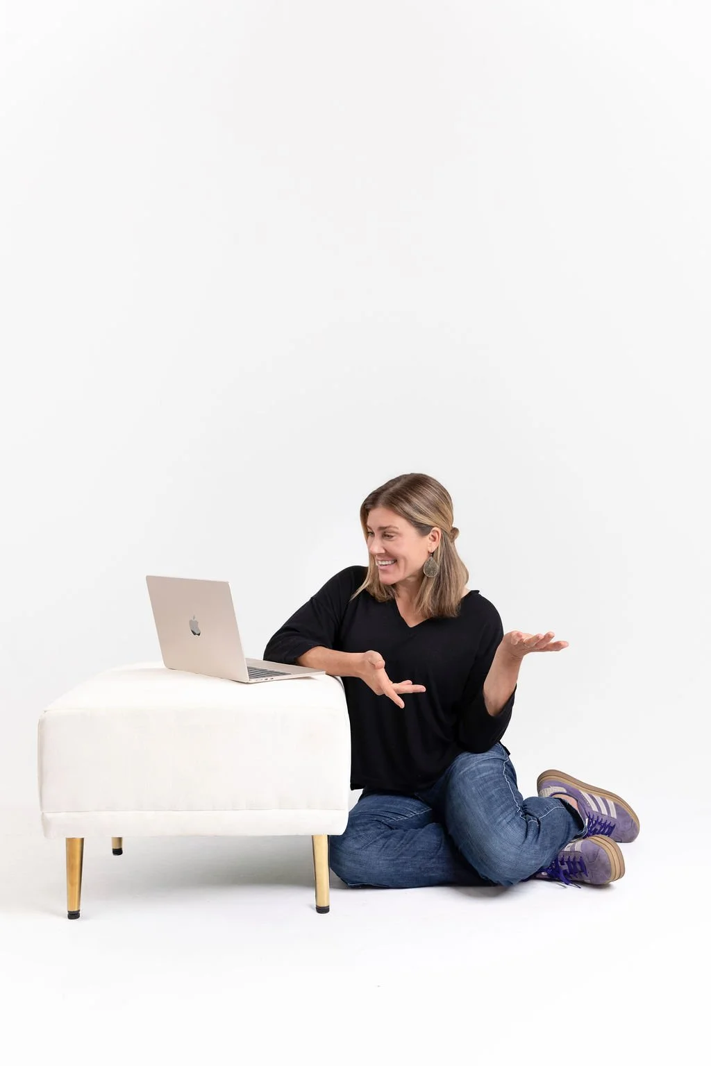 A woman sitting on the floor leaning on a white ottoman with a silver laptop, smiling and gesturing with her hands against a plain white background.