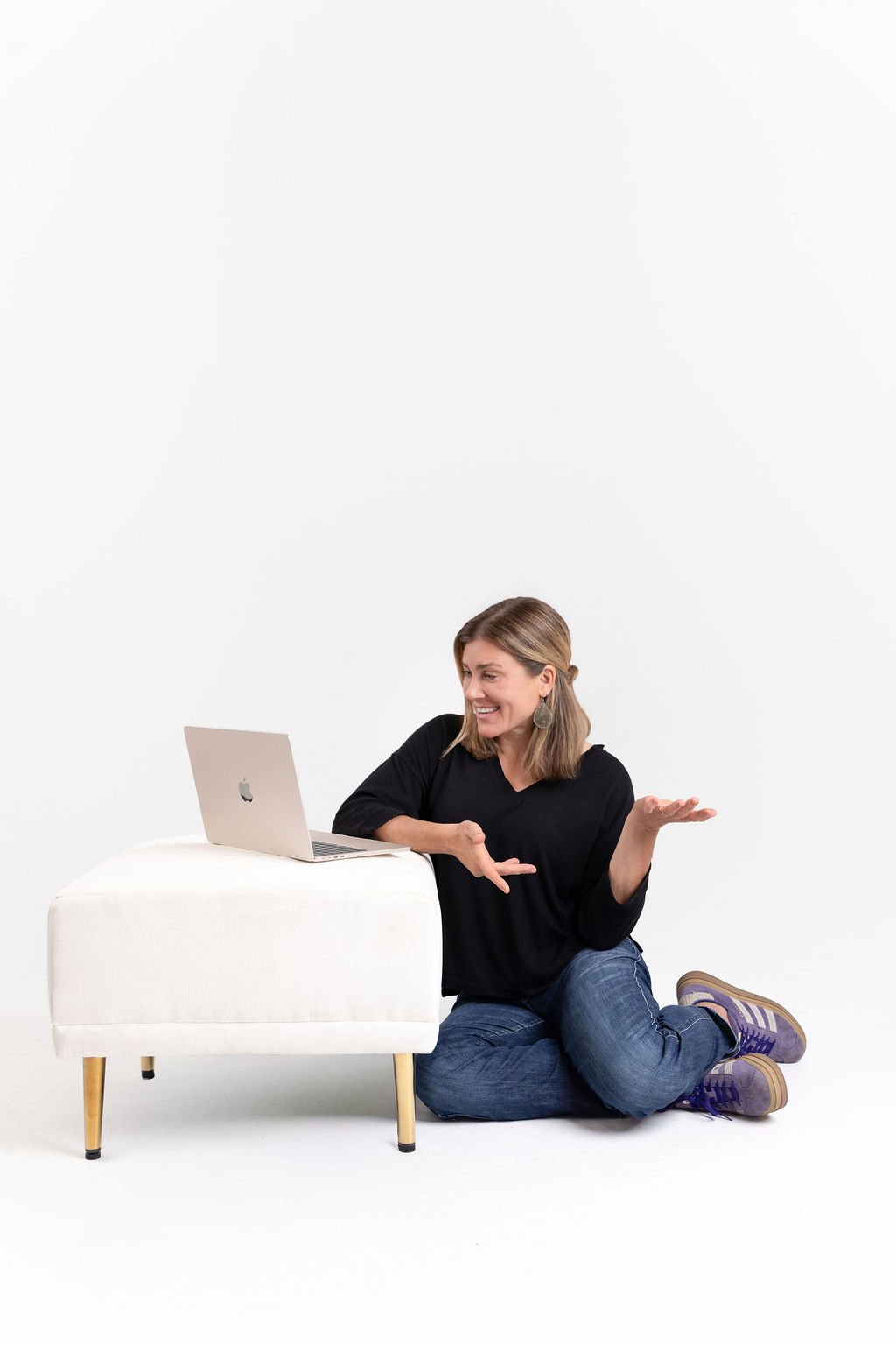 A woman sits on the floor with a white ottoman, a silver laptop on it, smiling and gesturing with her hands against a plain white background.