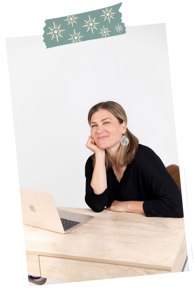 A woman sitting at a desk with a laptop, resting her chin on her hand and smiling at the camera.