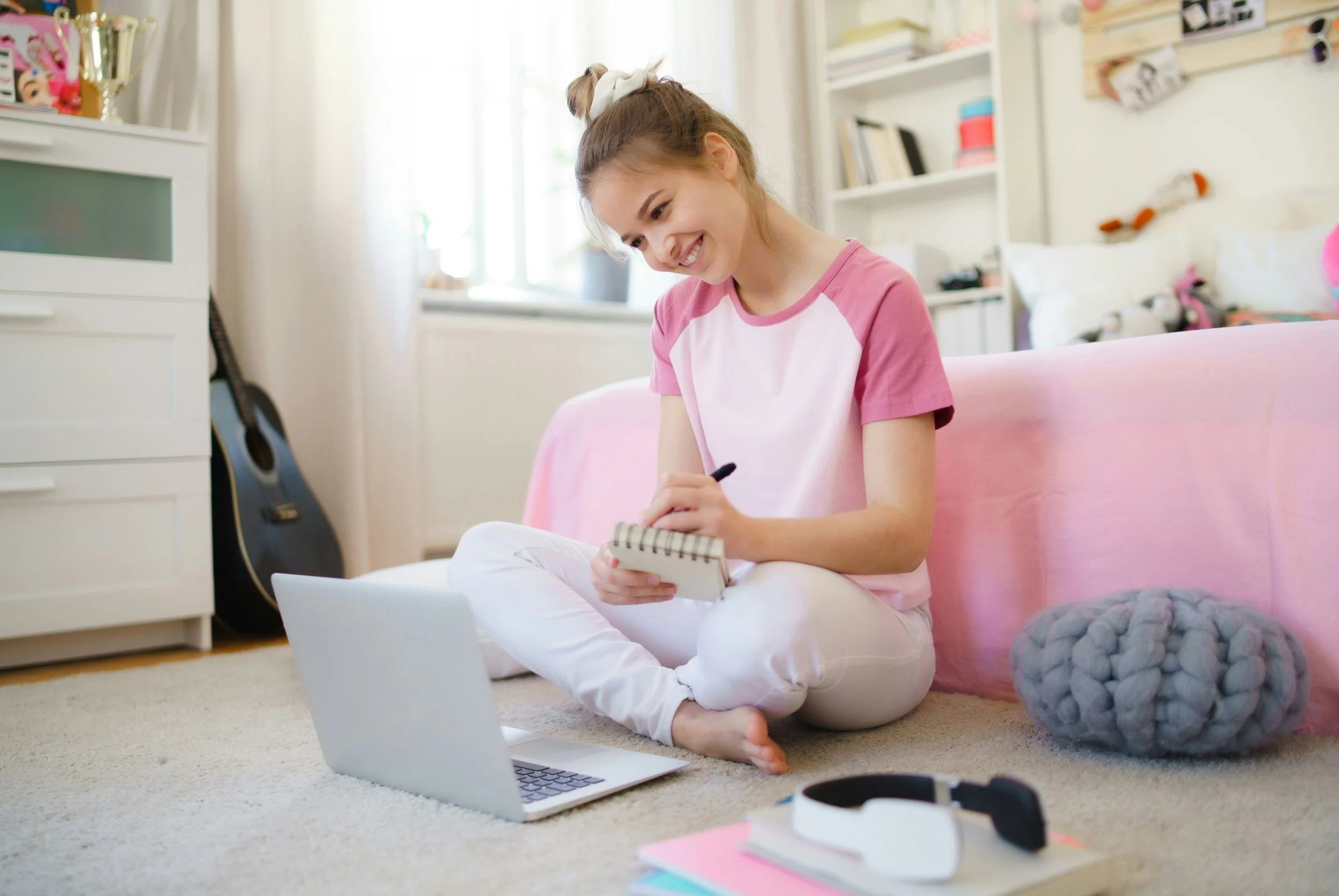 A girl sitting on the carpeted floor in a cozy room, smiling as she writes in a notebook, with a laptop open in front of her and various items, including a gray knit cushion, scattered around.