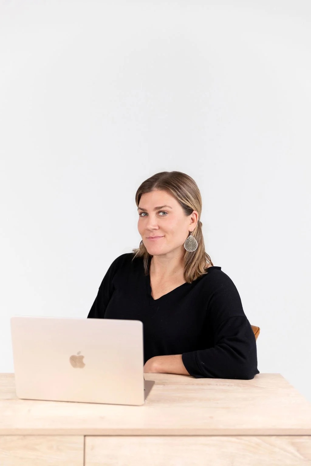 A woman with shoulder-length brown hair and large dangling earrings sitting at a light wooden desk with a closed silver MacBook.