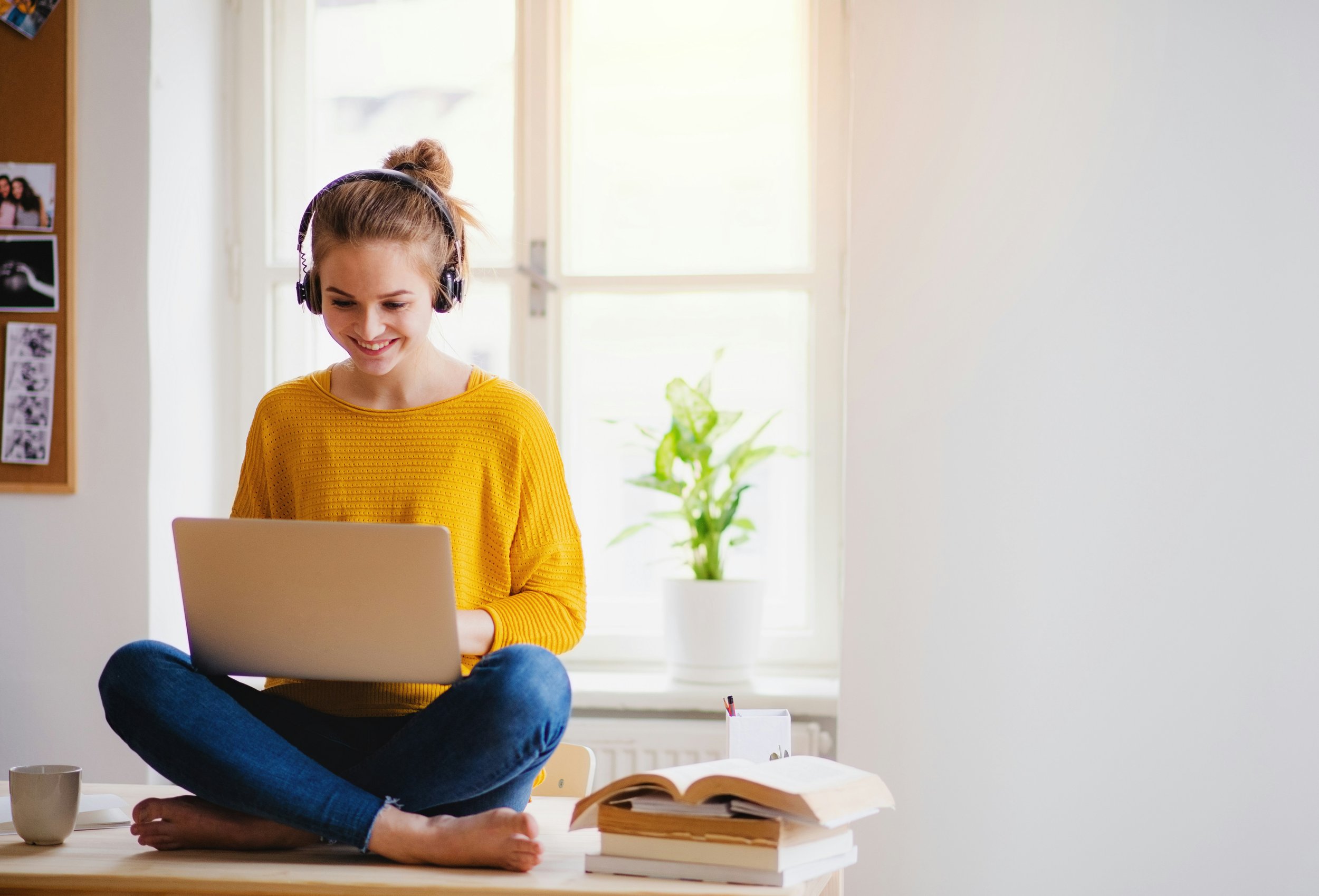 A young woman with light skin and blonde hair tied in a bun, wearing a yellow sweater and blue jeans, is sitting cross-legged on a table. She is smiling and looking at a laptop in front of her. She is also wearing large headphones. There are several books, an open notebook, a cup, and a pen holder on the table. The background shows a bright room with large windows, a potted plant, and photos on a corkboard.