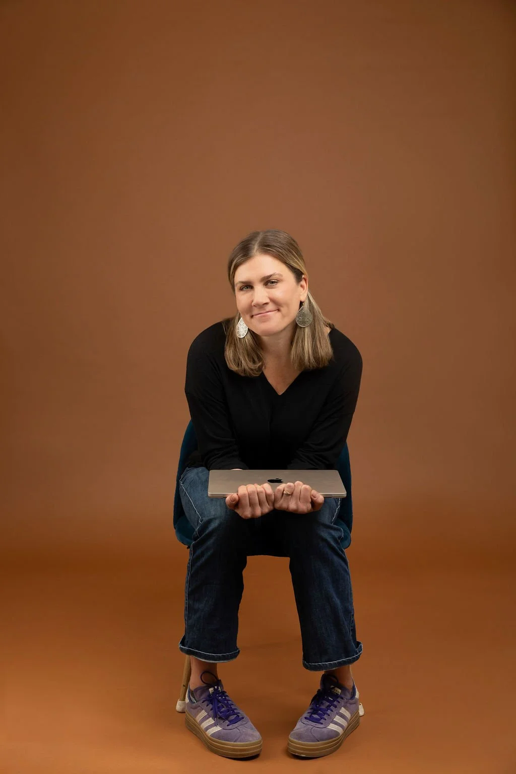A woman sitting on a chair, holding a closed silver laptop, smiling, wearing a black long sleeve top, blue jeans, purple sneakers with beige soles, and large silver earrings, against a plain brown background.