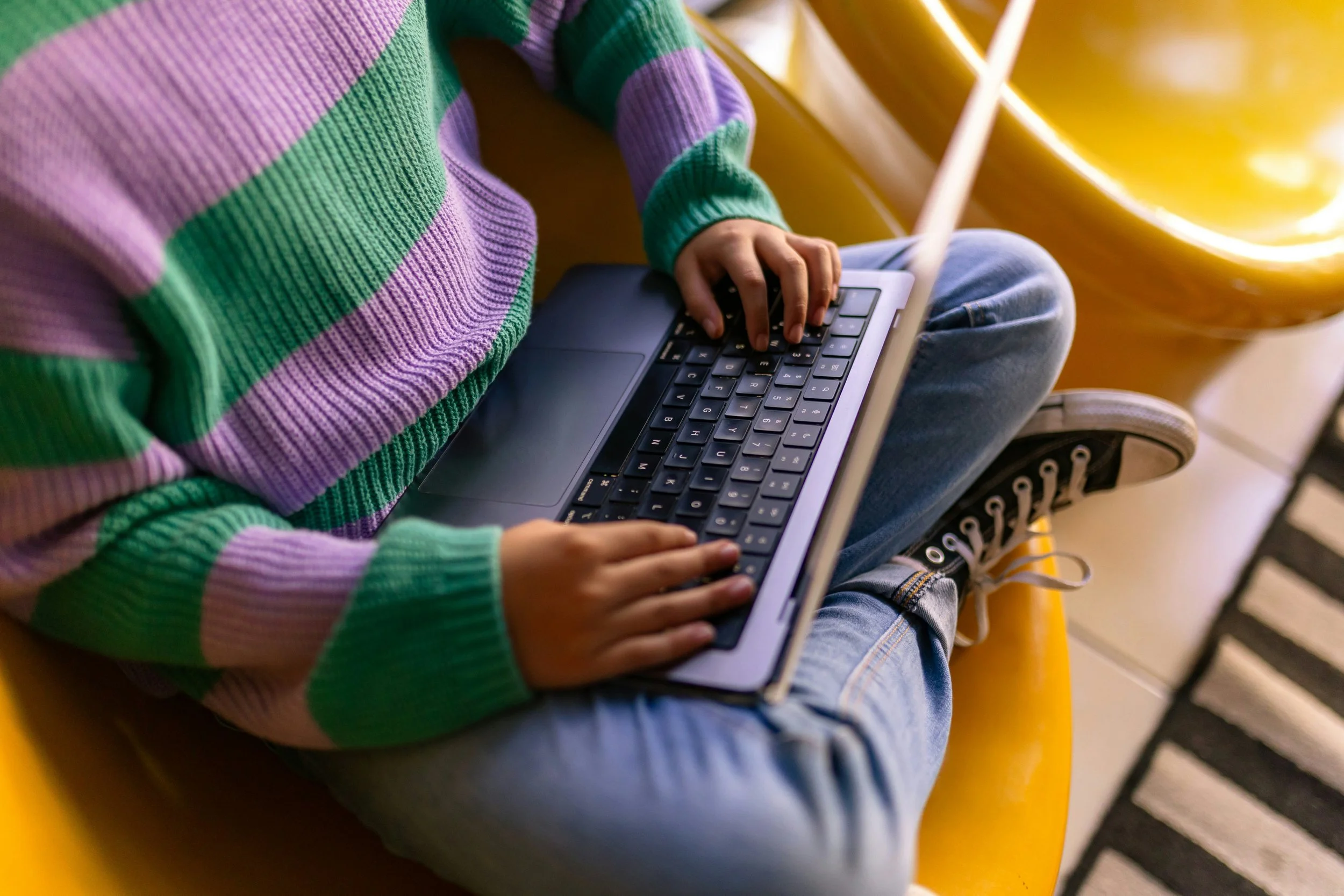 Teen doing online therapy using a laptop, wearing a colorful striped sweater and jeans, with a yellow playground slide in the background.