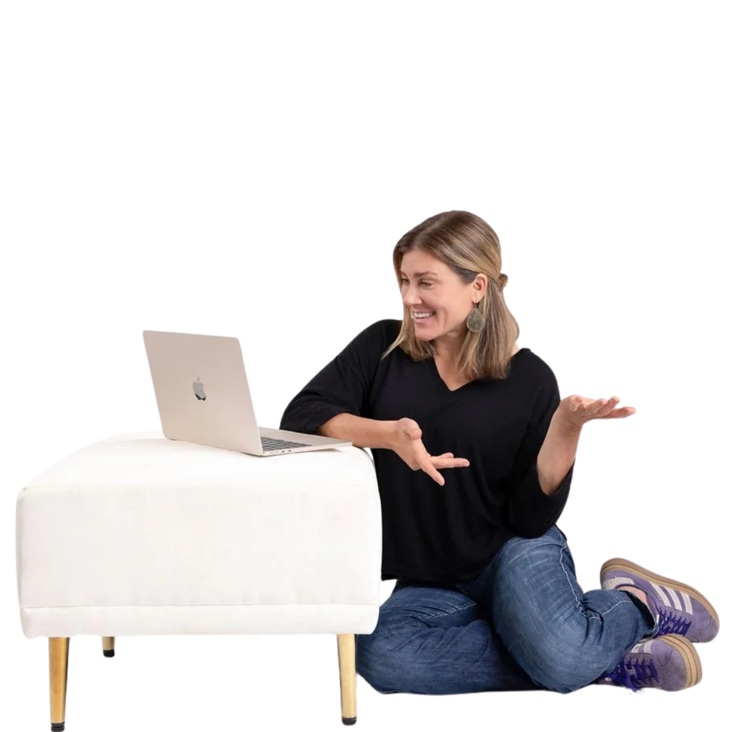 A woman with shoulder-length brown hair, wearing a black long-sleeve shirt and jeans, sits on the floor with her legs crossed. She has a notebook and an open MacBook on a white, modern-style table in front of her. She is smiling and gesturing with her hands while looking at the laptop.