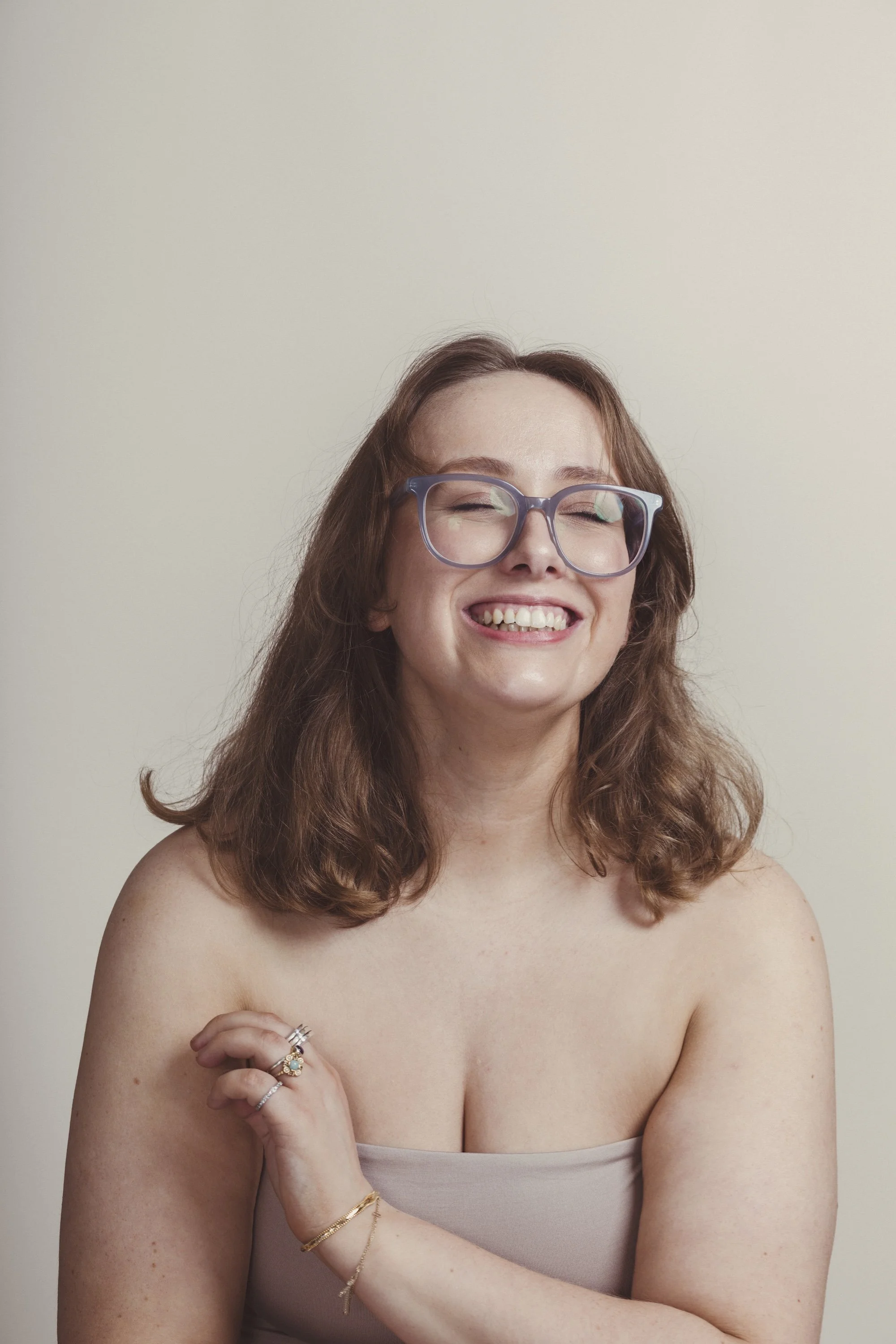A woman with wavy, shoulder-length reddish-brown hair, wearing large purple glasses, smiling with her eyes closed, with multiple rings on her left hand, and jewelry on her right wrist and left wrist, partially nude with a strapless top, against a pla