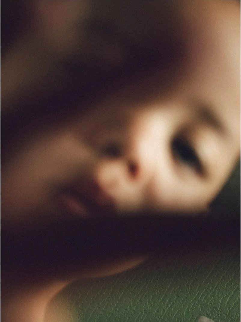 Close-up of a person's face, blurry, with dark hair and light skin, resting on a dark surface.