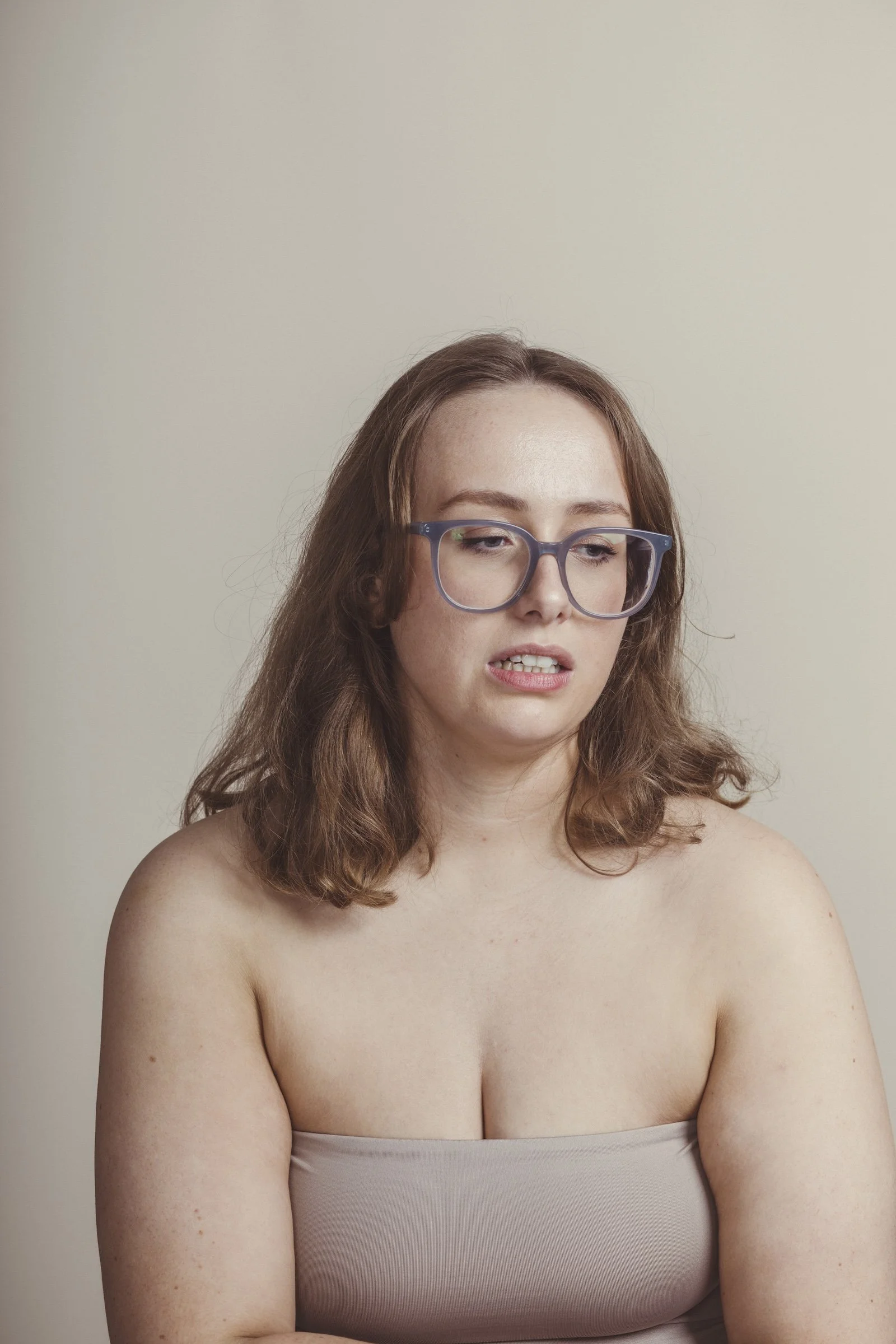 A woman with long, wavy auburn hair, wearing large purple glasses and minimal clothing, is looking down with a contemplative expression against a neutral background.