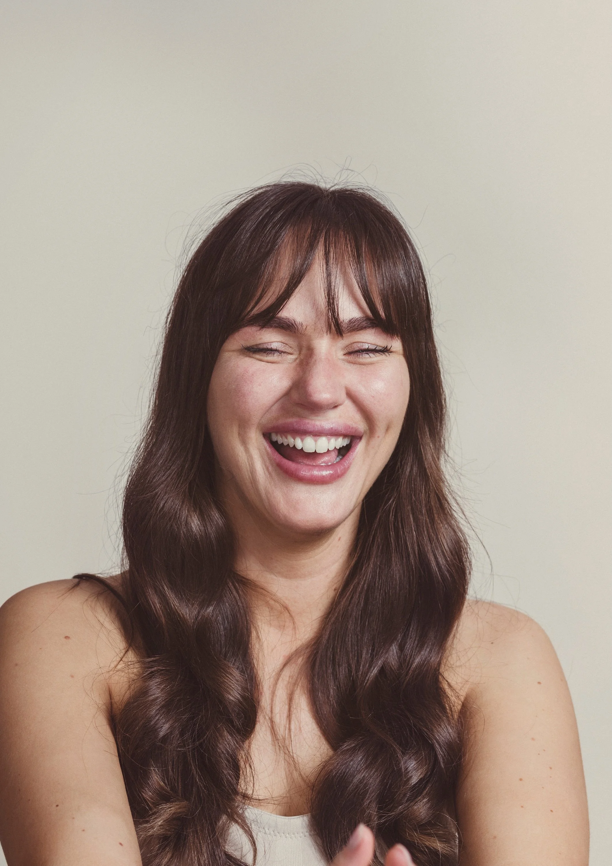 A woman with brown, wavy hair and bangs smiling broadly with her eyes squinting, against a plain, light-colored background.
