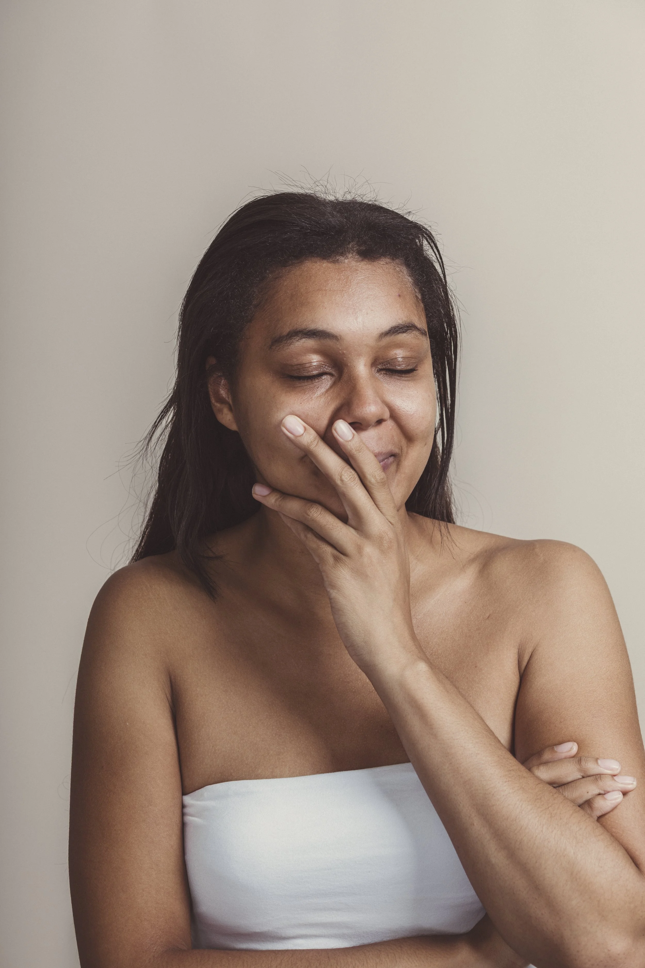 A woman with medium skin tone and dark hair, wearing a white strapless top, with her eyes closed, touching her face and chin with her right hand, in front of a plain light beige background.
