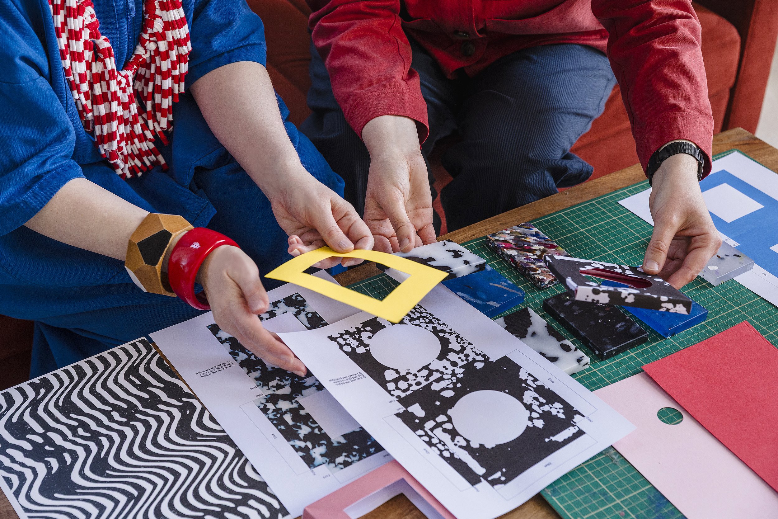 Two people working on a paper crafting project with black and white patterned papers, yellow paper frames, and various cutouts on a green grid cutting mat on a wooden table.