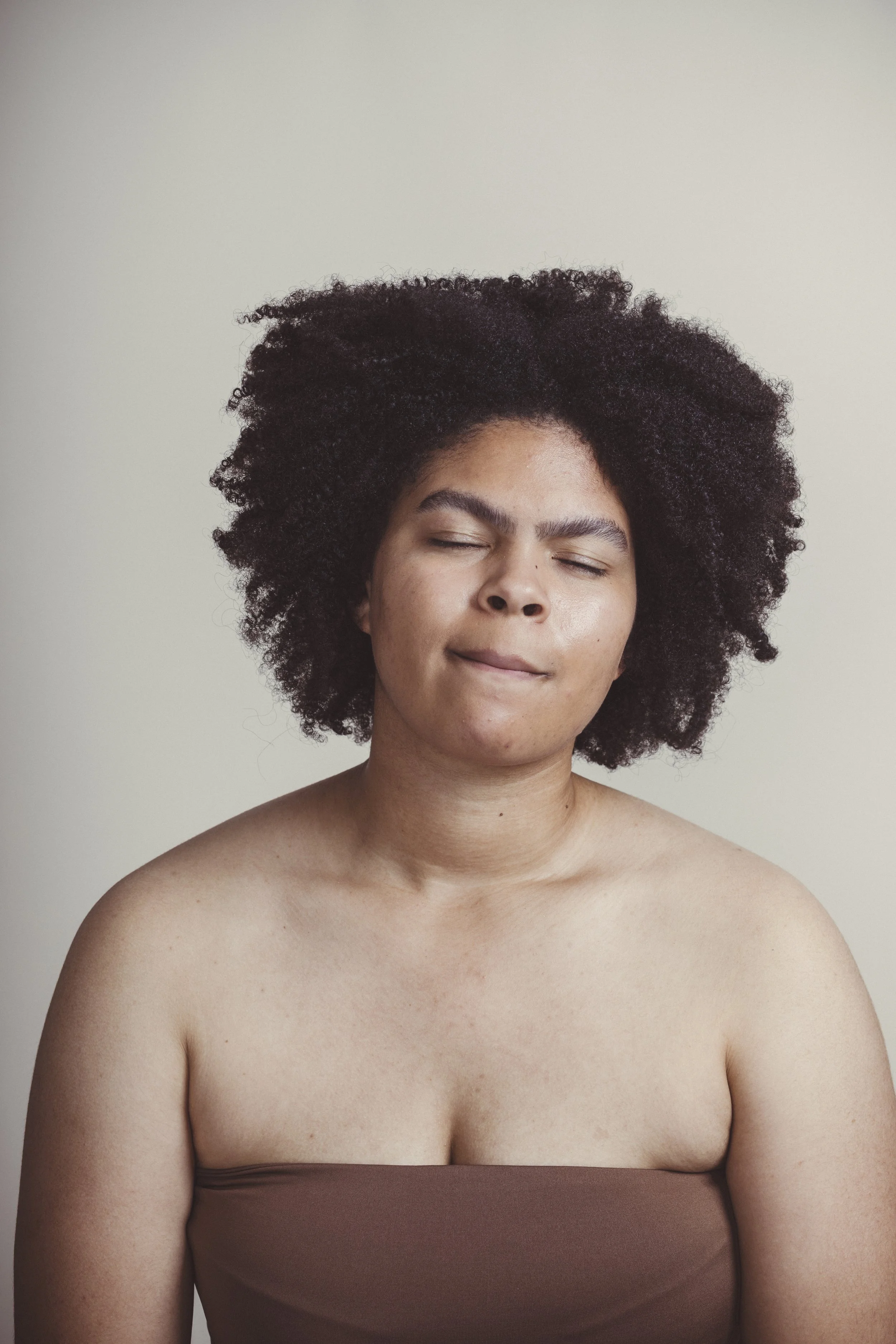 Portrait of a woman with closed eyes, natural curly hair, and bare shoulders, against a plain light background.