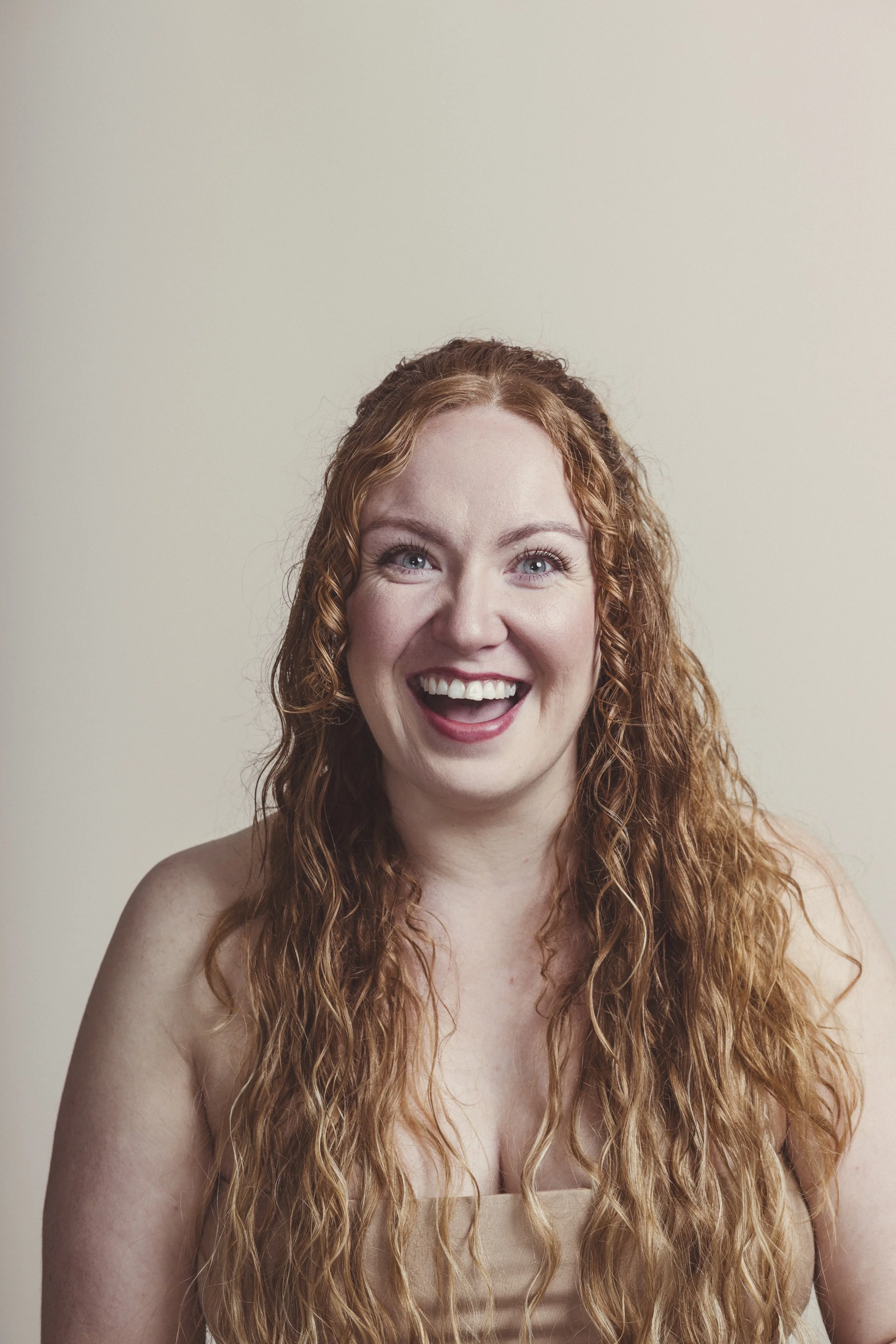 A woman with long, curly red hair smiling cheerfully, wearing a beige strapless top against a plain light background.