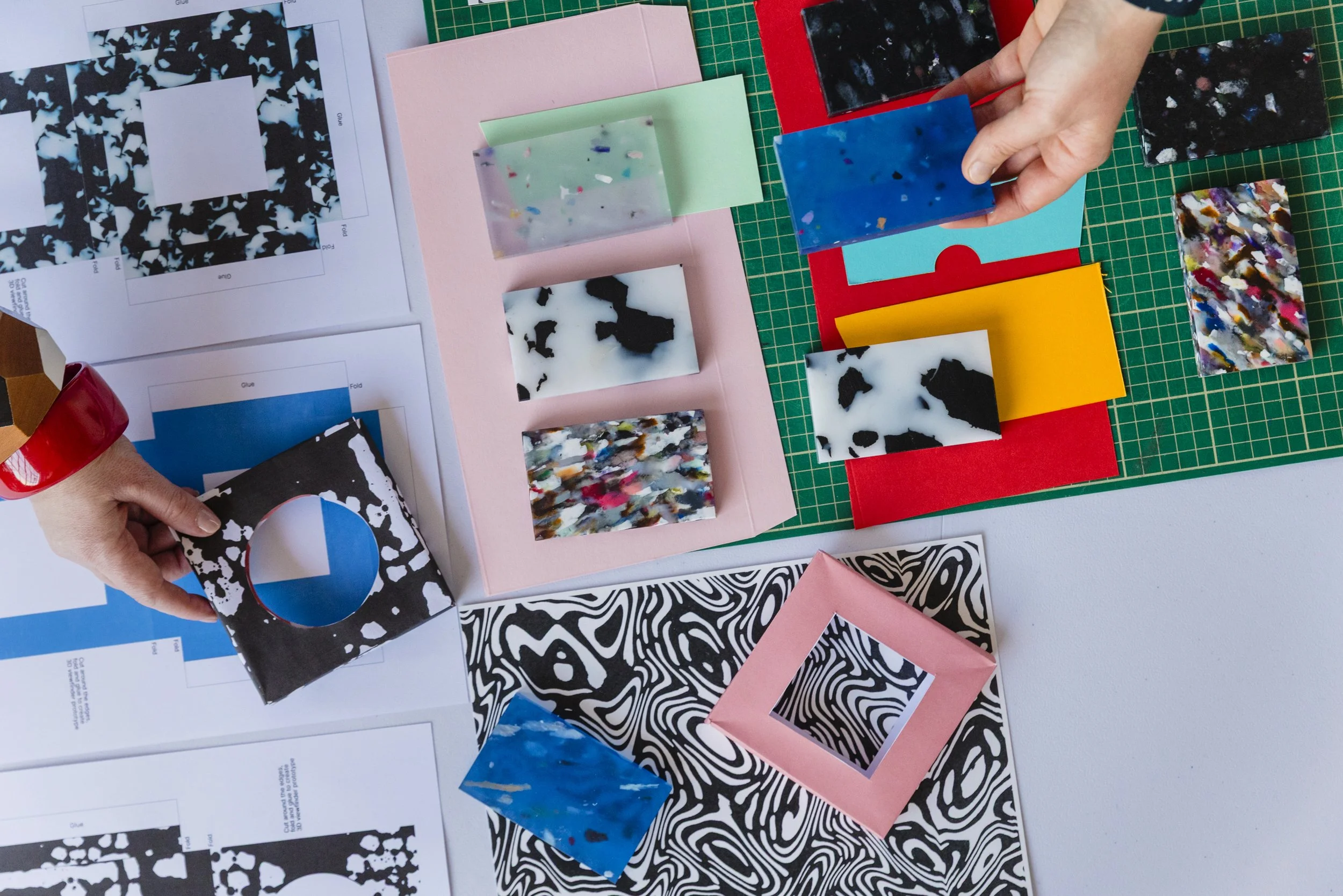 A person is working on a craft project involving various colorful acrylic or resin sheets with marbled, black and white, and solid colors. The workspace is covered with printed guides and sample pieces, with some hands holding and arranging the sheet