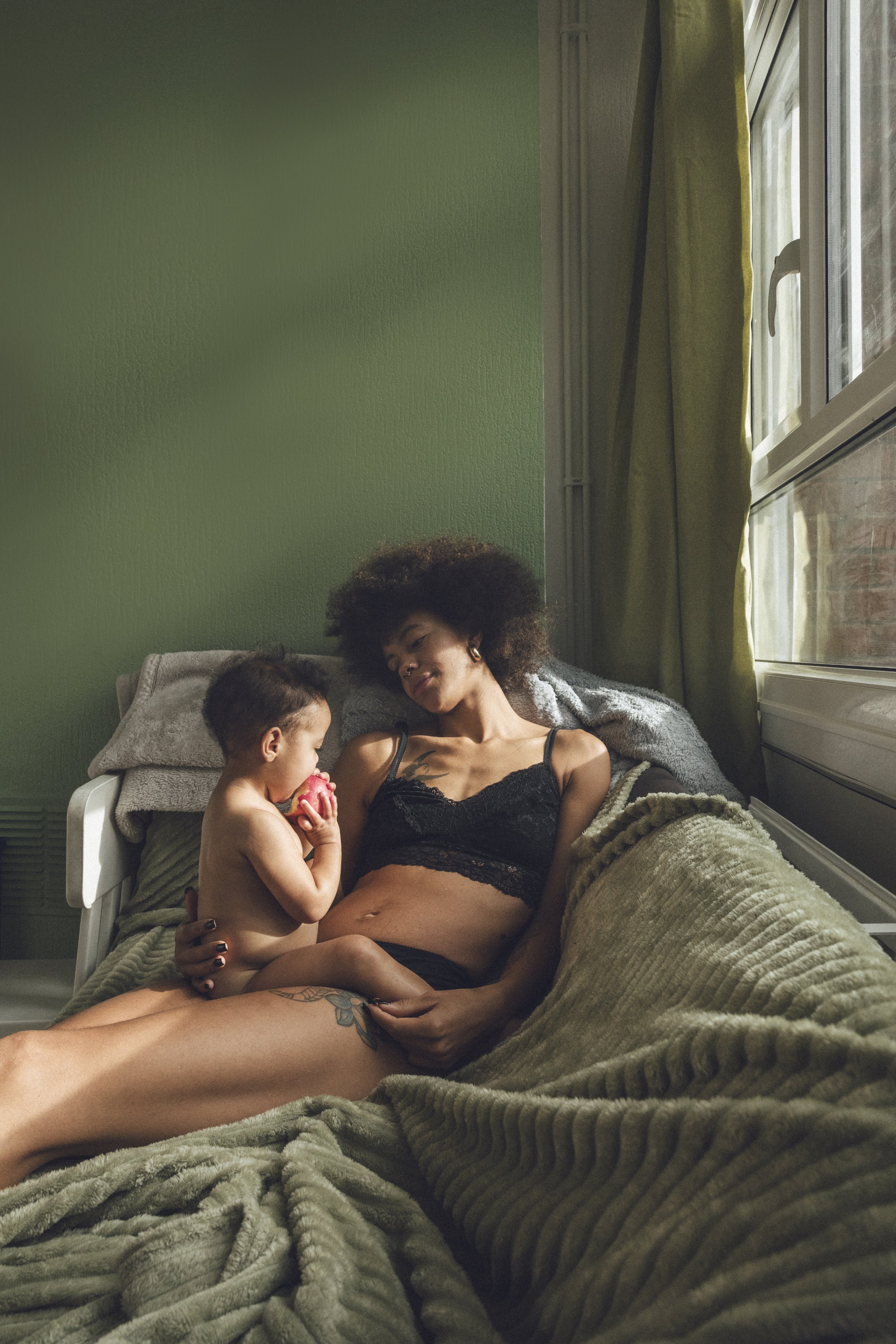 A woman and a young child sitting on a bed in a bedroom. The woman is lying back with her head against a pillow, wearing a black lace tank top, and the child is sitting on her lap, holding a pink toy and looking at her. The room has green walls, a la