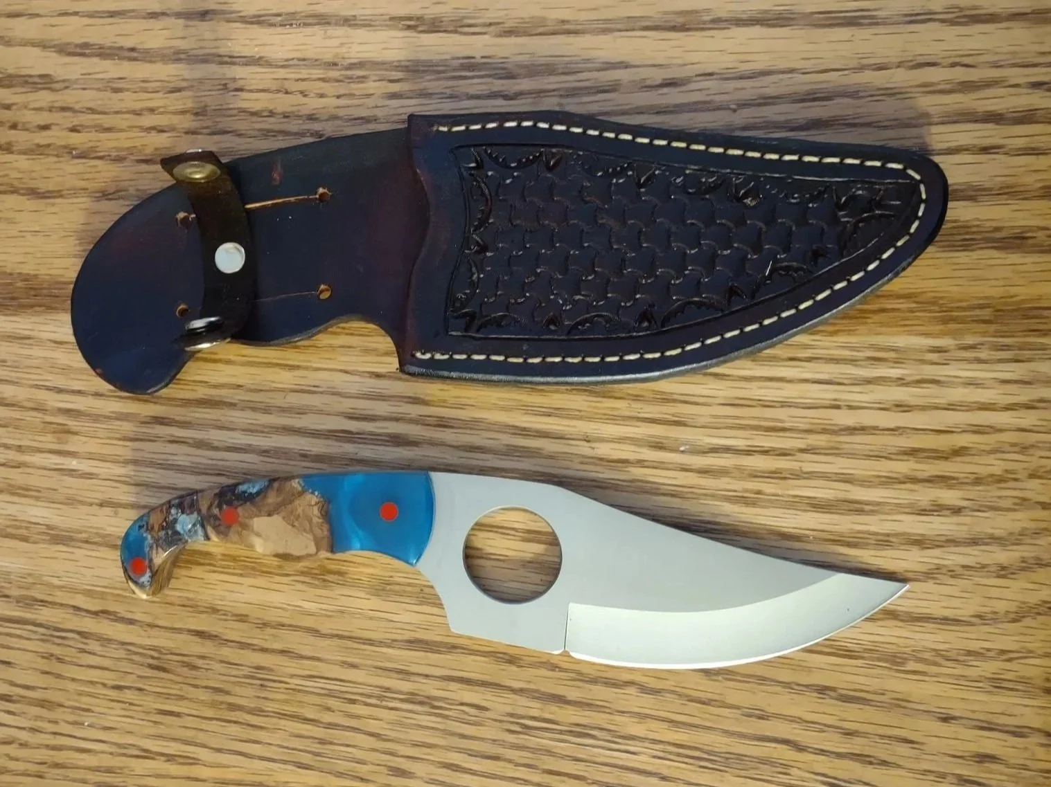 A small skinner knife with a curved blade and a hole near the handle, alongside a black leather sheath, both placed on a wooden surface.
