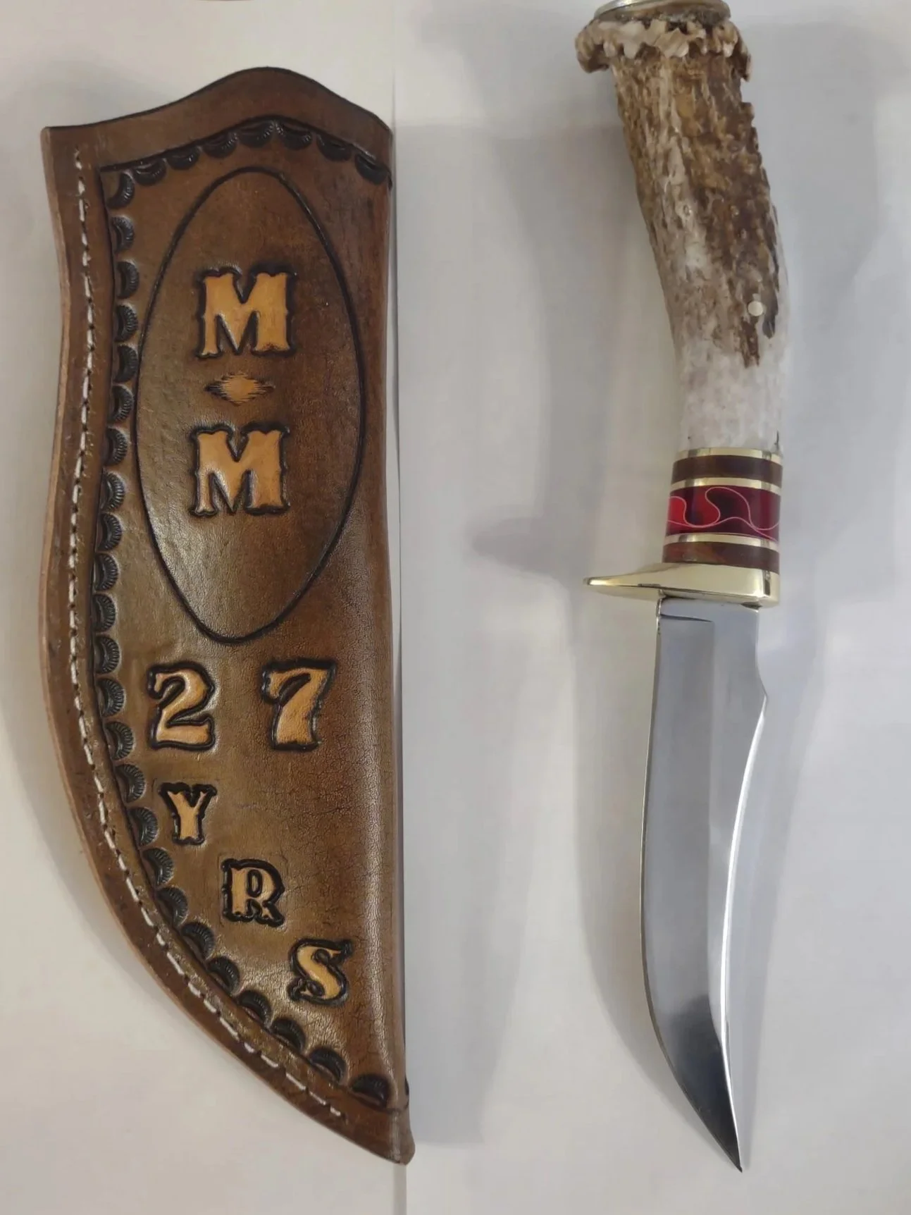 A hunting knife with a decorative deer antler handle and a brown leather sheath with embossed lettering and a stitched border.
