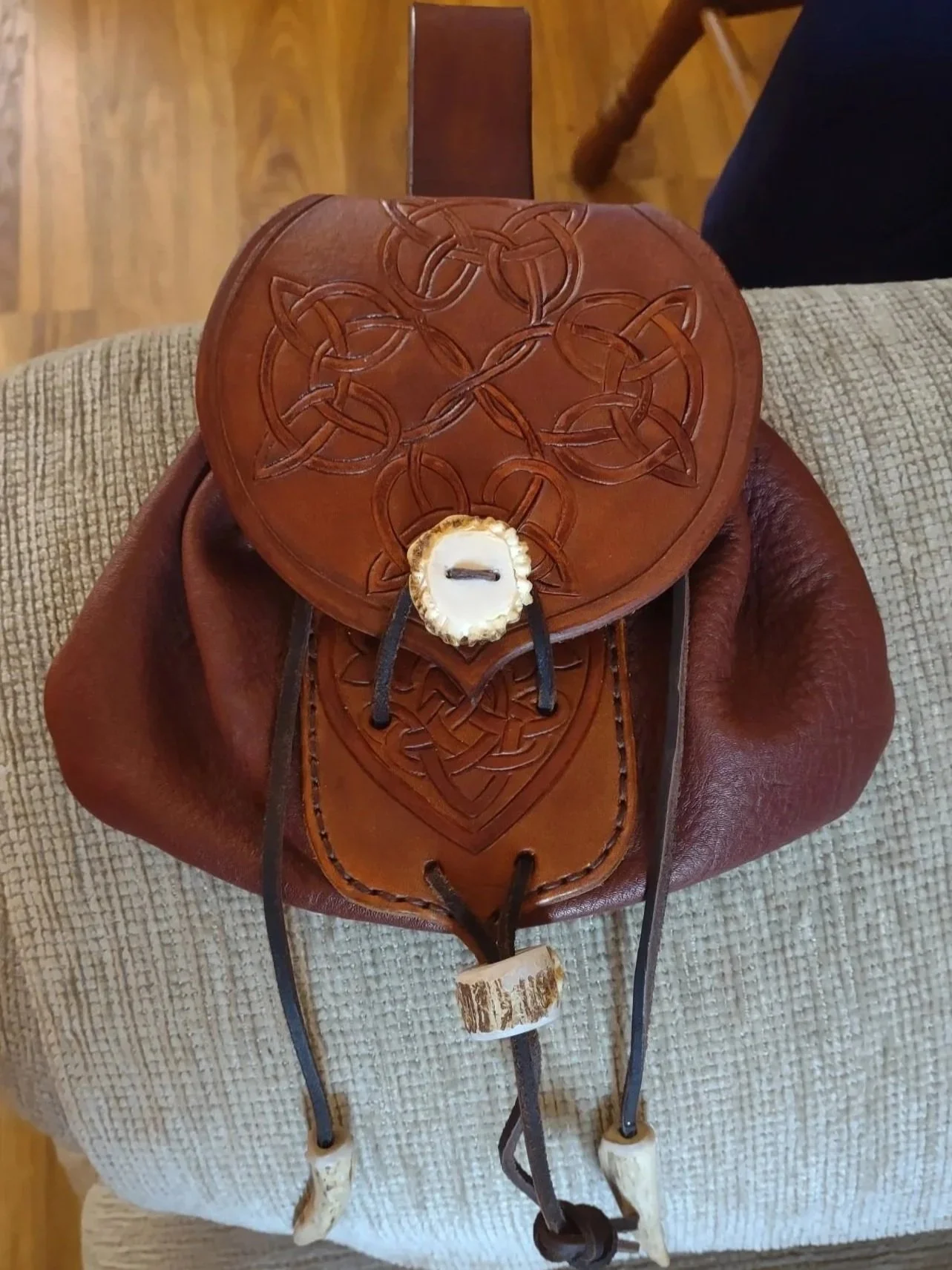 A leather satchel with decorative carvings and hand stitching placed on a beige textured blanket. The satchel has leather tassels and a leather flap and button closure at the front.