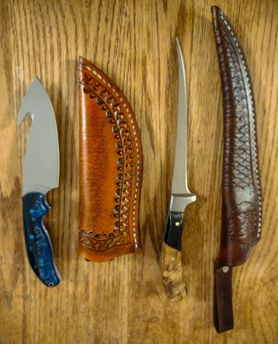 Two custom, handmade knives with sheaths lying on a wooden surface. The first knife has a blue handle and a decorative leather sheath. The second knife has a black and wooden handle with a handmade leather sheath.