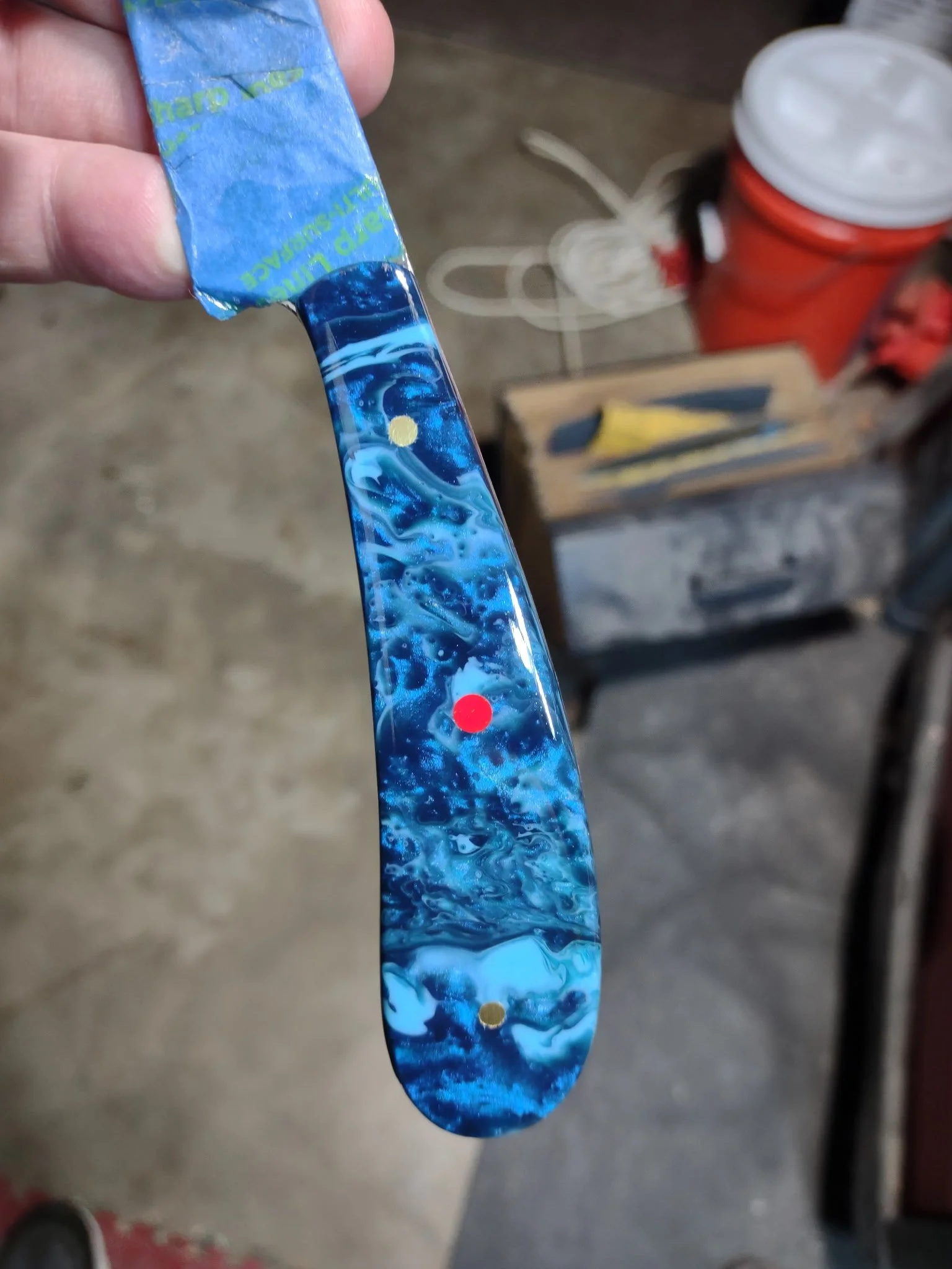 A person holding a blue and white marbled knife handle with a yellow red pins, in a workshop setting with a wooden surface in the background.