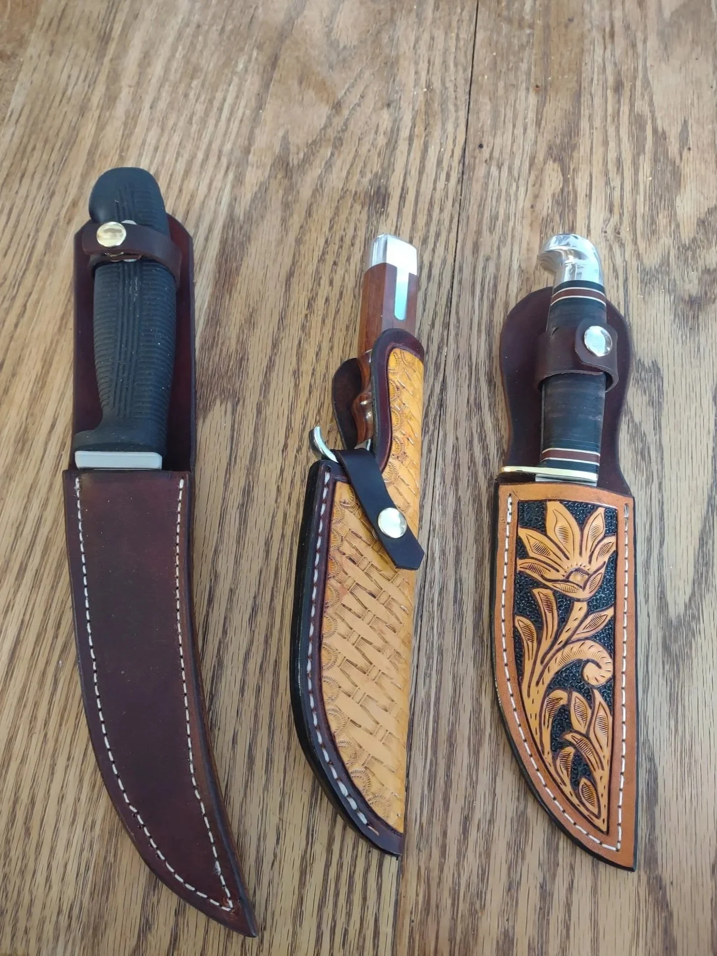 Three custom made knives placed in decorative leather knife sheaths placed on a wooden surface.