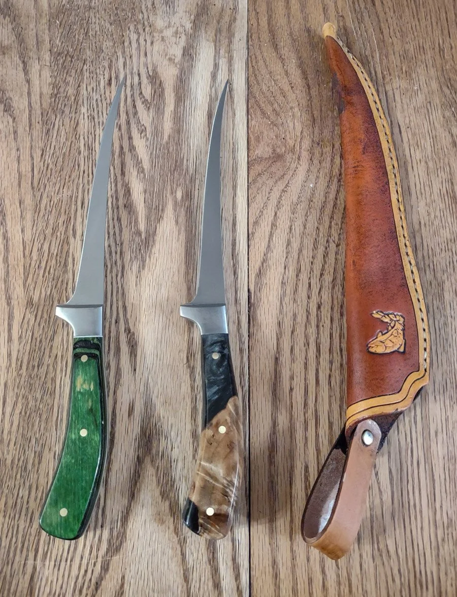 Two custom, hand made fillet knives with different colored handles and a custom, hand made leather sheath on a wooden surface.