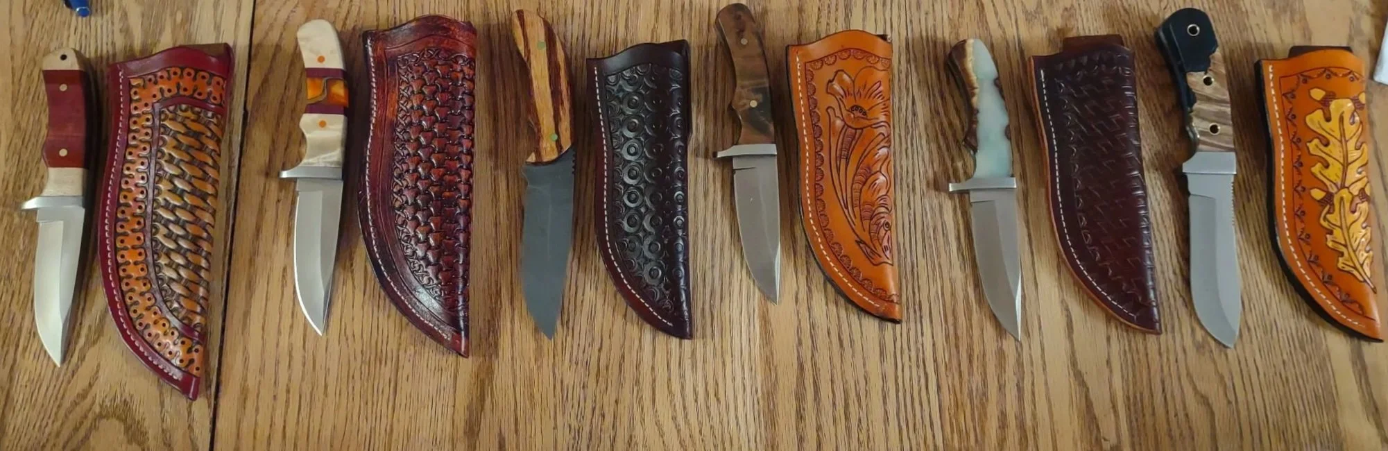 A row of ten decorative custom made knives  and sheaths. The knives have various ornate handles, laid out on a wooden surface. Each knife has a distinct handle design, with some featuring intricate carvings and colorful patterns.