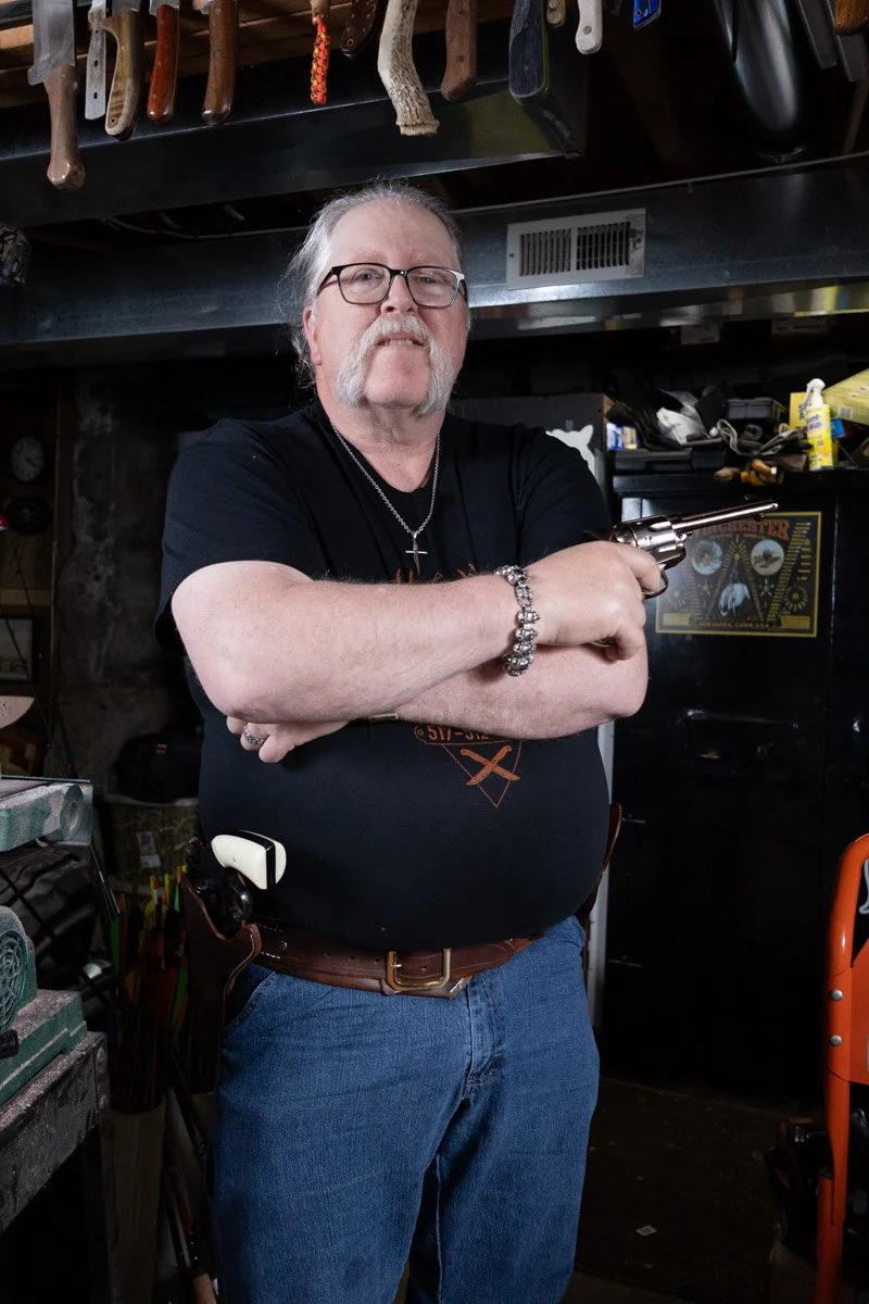 A man with glasses, a mustache, and a goatee, holding a revolver, standing in a workshop with tools hanging above him and various equipment around him.