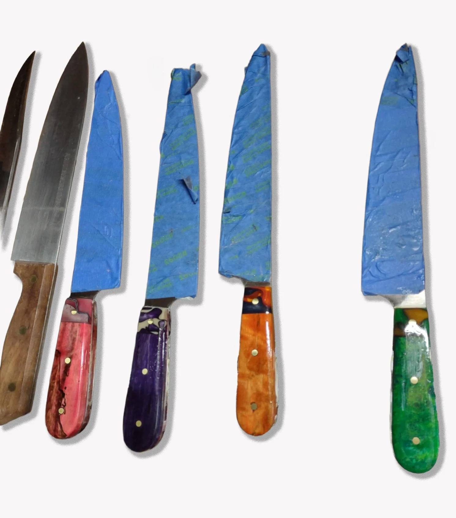 Six knives with colorful, patterned handles and blue taped blades for production purposes.