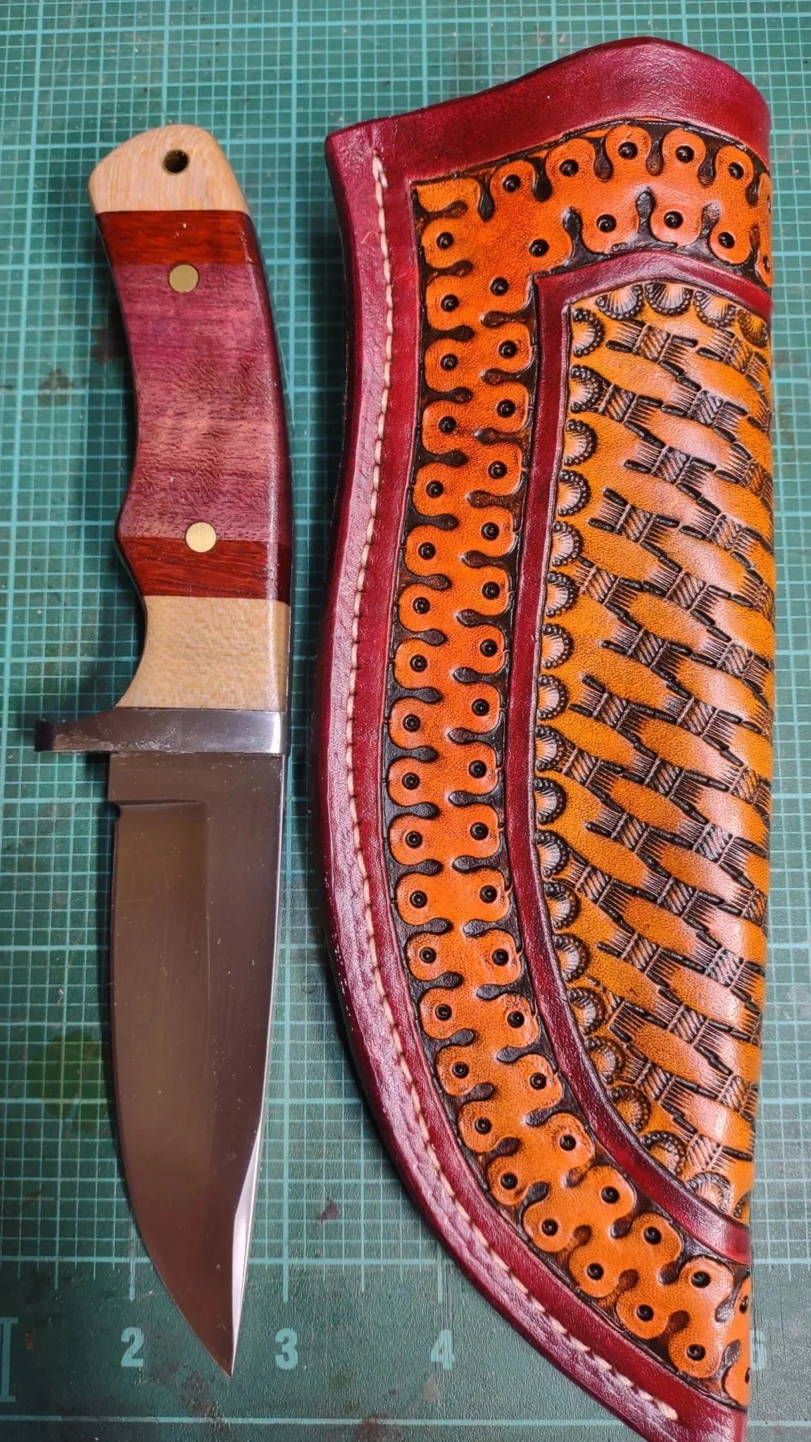 A fixed-blade knife with a wooden handle resting on a green cutting mat with measurements, next to a colorful, decorative leather sheath.