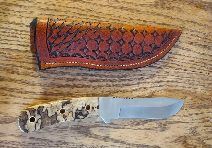 A custom, handmade fixed-blade hunting (skinner) knife with a textured, patterned handle and a sharpened blade, placed below a decorative leather sheath with embossed geometric designs.