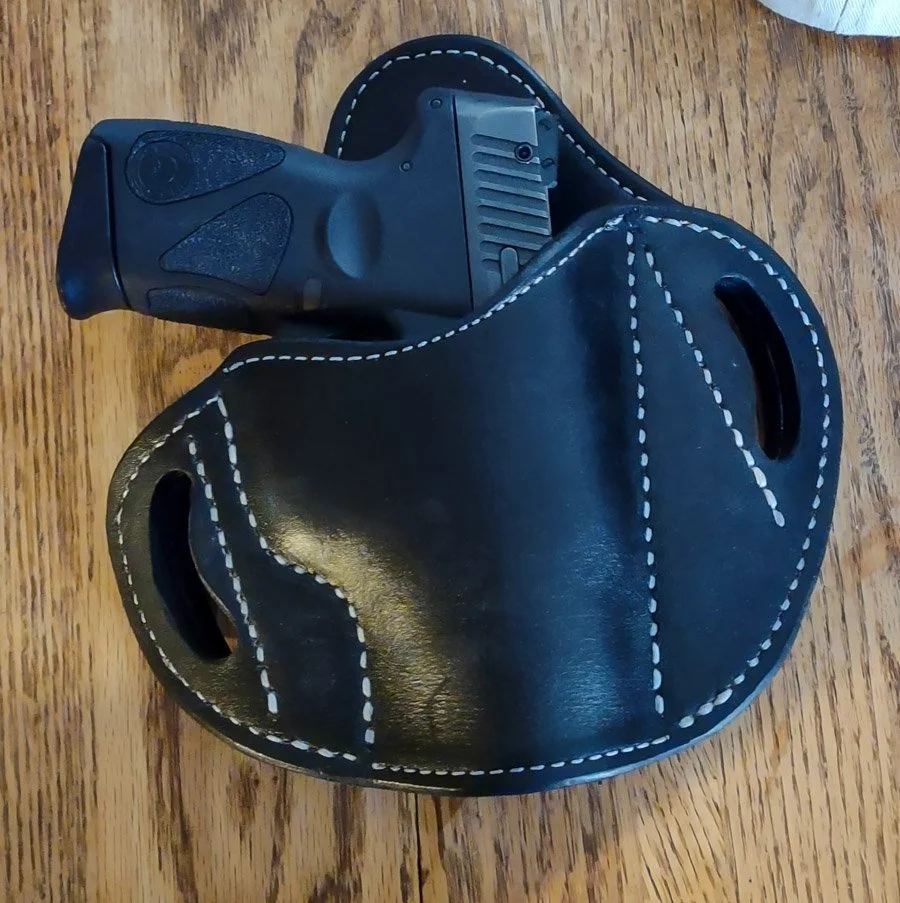 A black handgun inside a black leather gun holster with white stitching on a wooden surface.