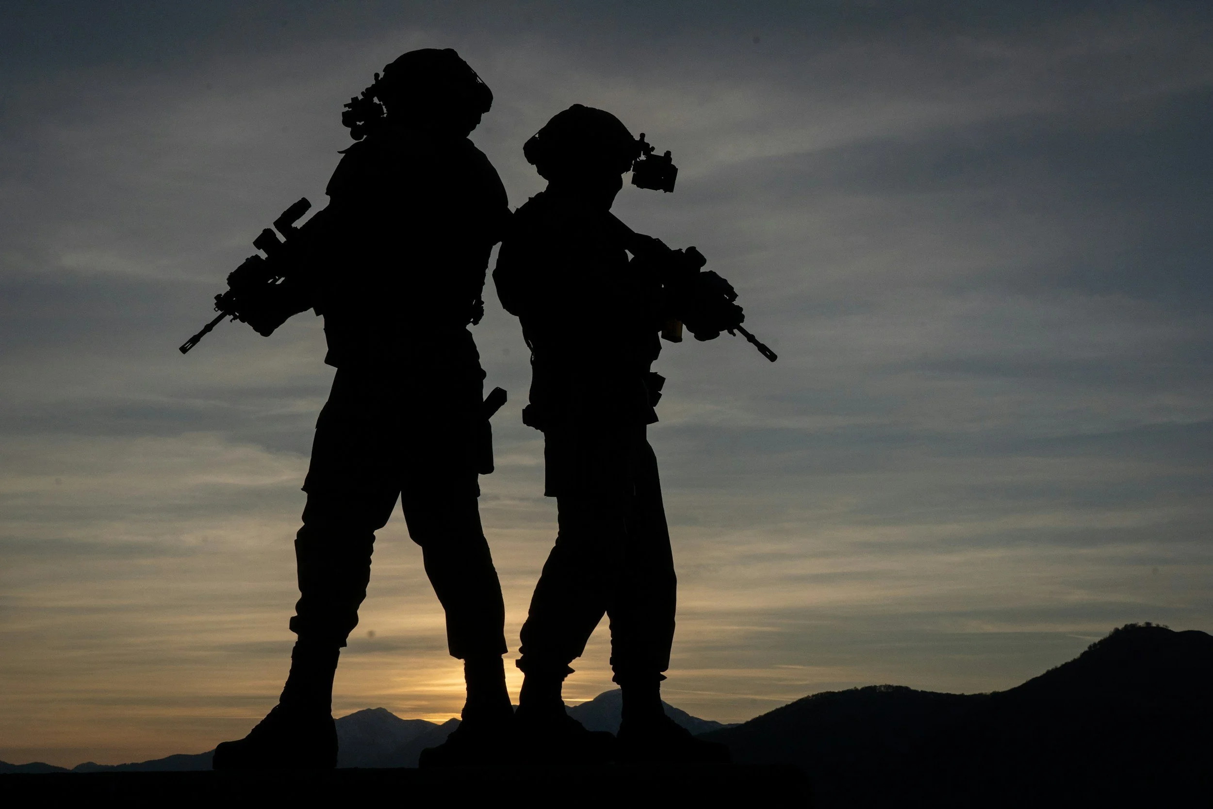 Silhouettes of two soldiers standing on a hill at sunset, with mountains and a cloudy sky in the background.