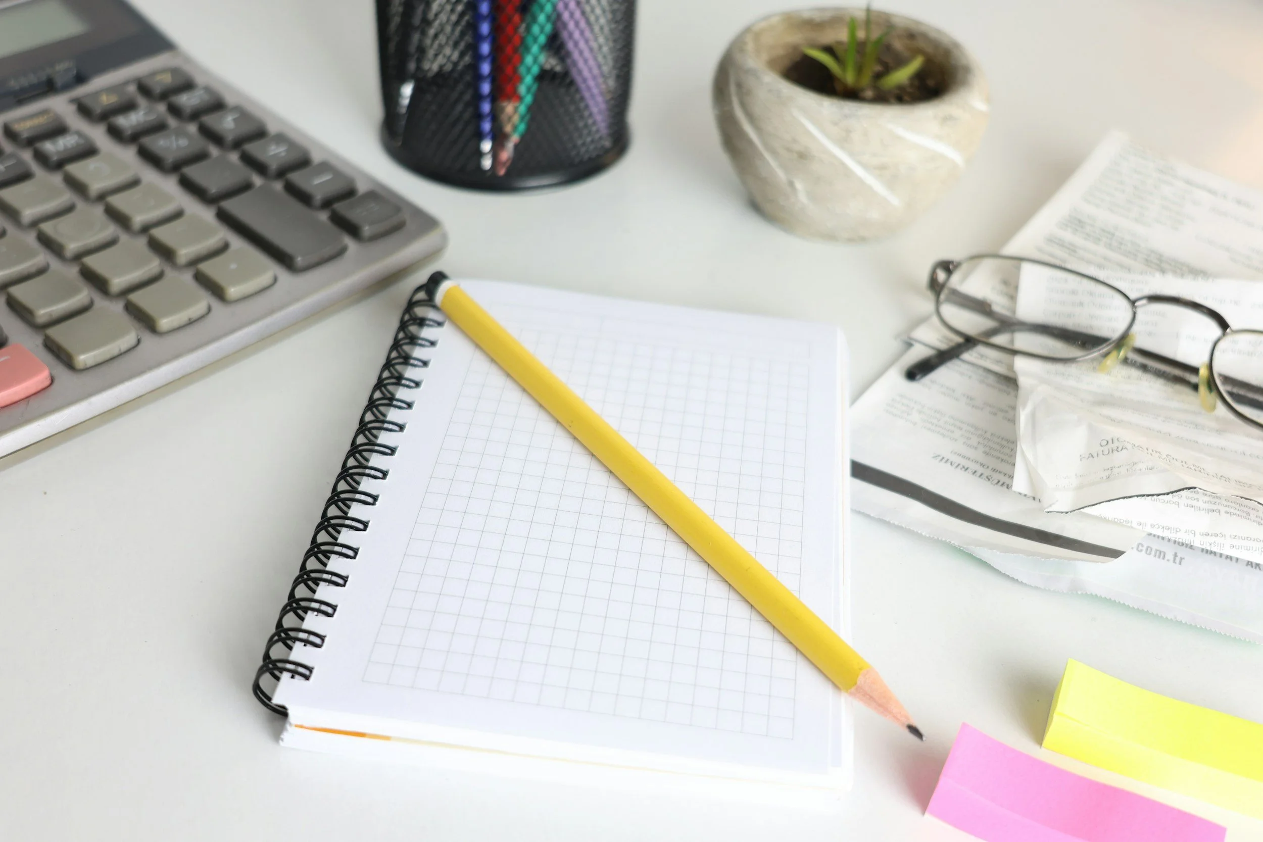 Desk workspace with a computer keyboard, a black pen holder with colored pens, a small potted plant, a pair of reading glasses, an open book, a notebook with a yellow pencil, and some sticky notes.