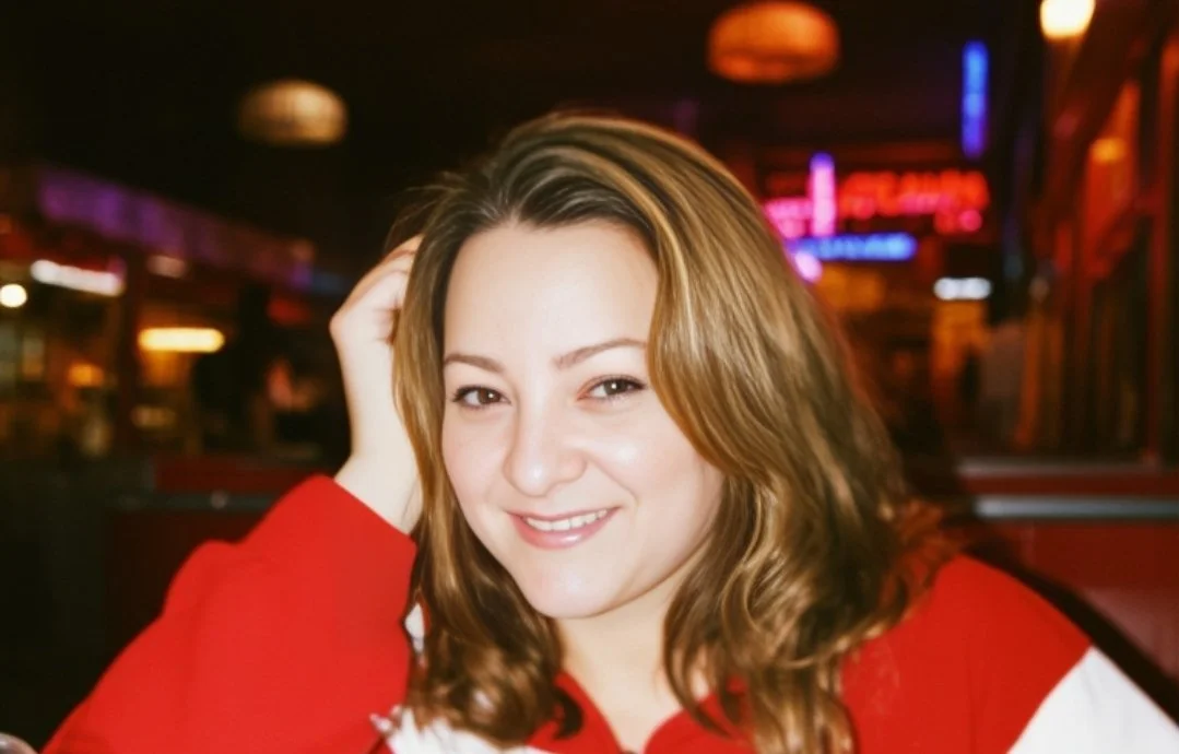 A smiling woman with shoulder-length light brown hair, wearing a red and white top, in a dimly lit, colorful bar or restaurant.