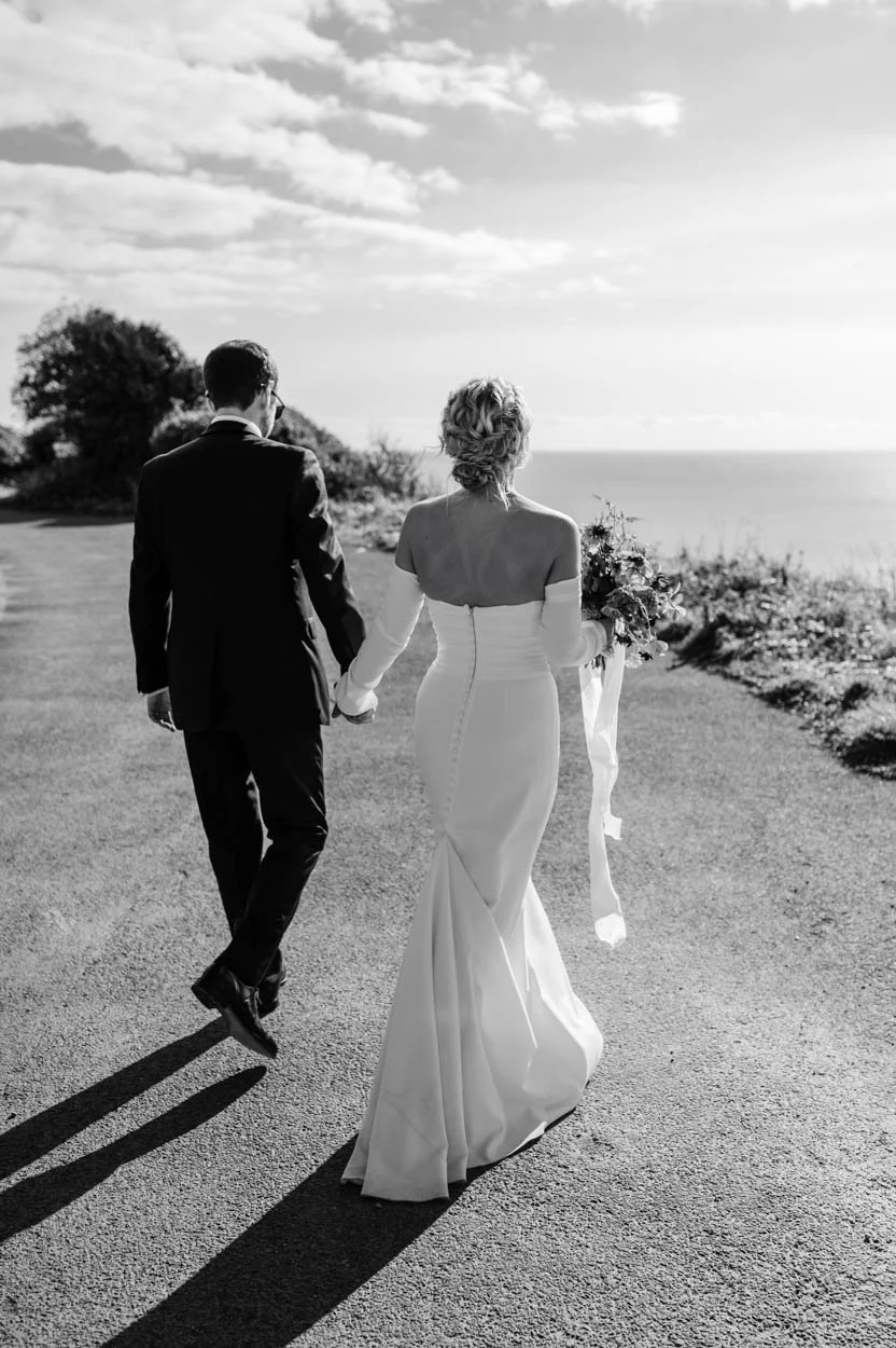A black and white photo of a bride and groom holding hands while walking outdoors near the Devon coast, with the ocean in the background and partly cloudy sky.