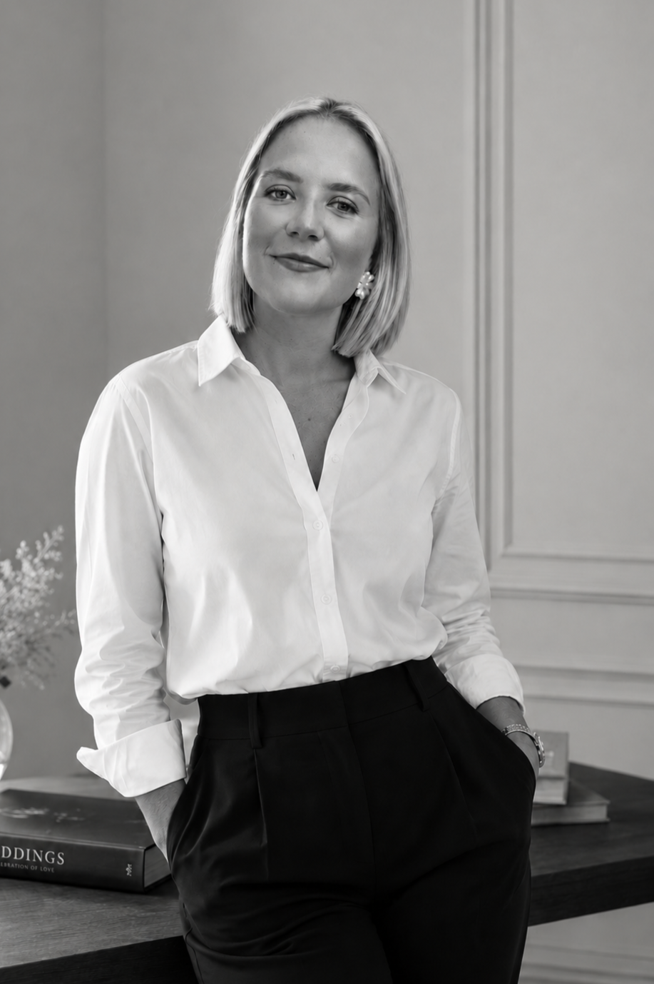 Devon Wedding Planner Simone Burchell wearing a white button-up shirt and black pants, standing in a room with a wooden desk and decorative molding on the wall, smiling at the camera.