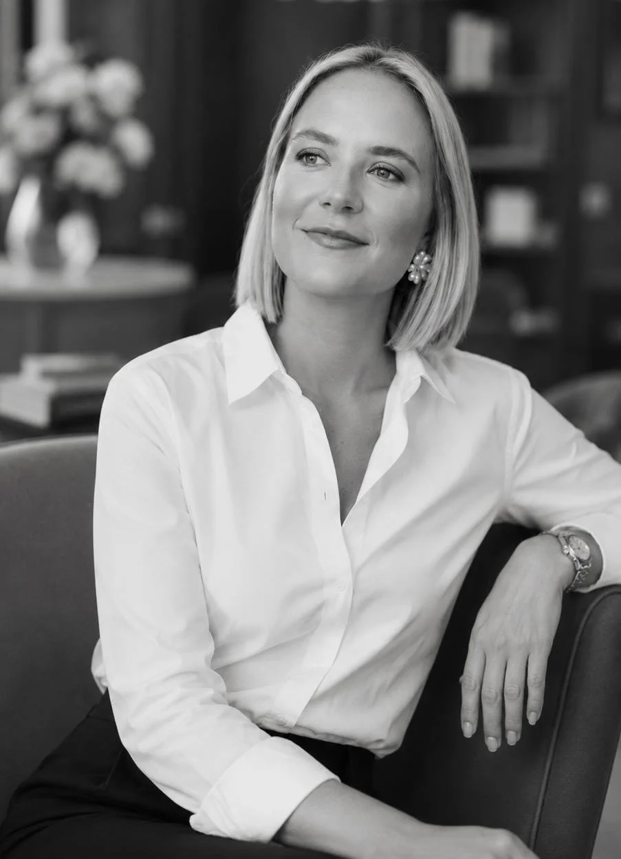 Black and white photo of Devon wedding planner Simone Burchell wearing a white button-up shirt and earrings, sitting on a chair in a room with bookshelves and a vase of flowers in the background.