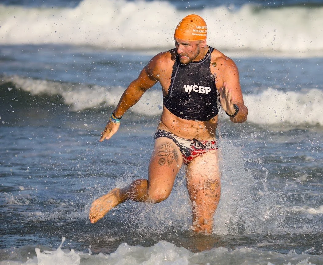 A man running out of the ocean wearing a wetsuit top, American flag shorts, and an orange swim cap.