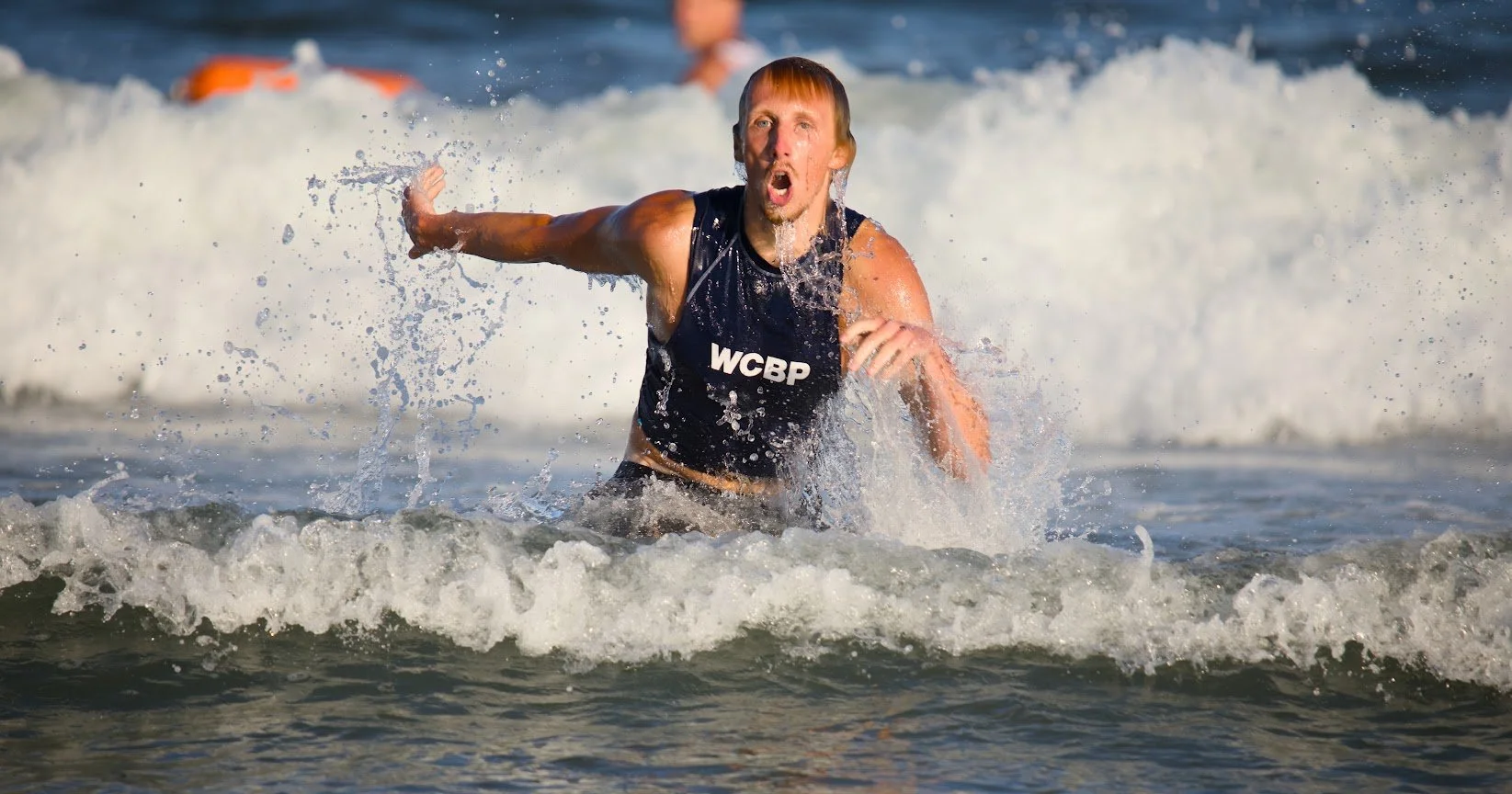 A man wearing a 'WCBP' shirt is running out of the ocean waves, looking surprised or excited