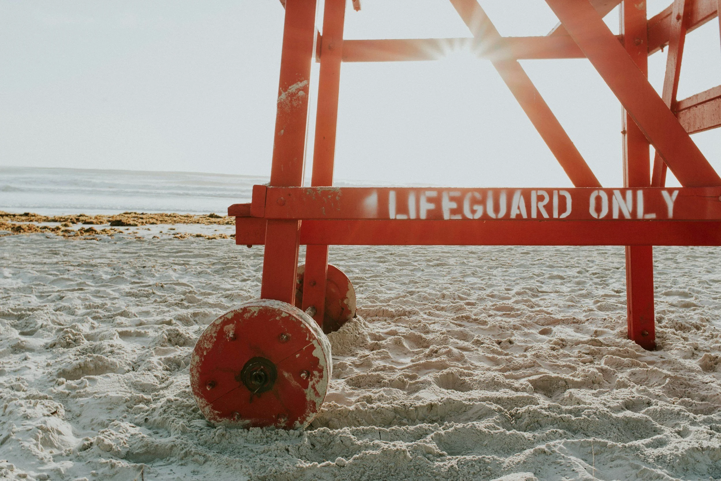 A red lifeguard tower on a sandy beach with ocean waves in the background, sunlight shining through the structure.