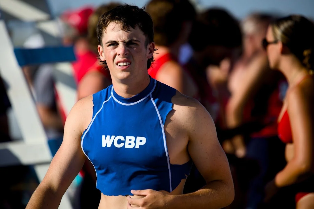 Young male athlete wearing a blue sleeveless sports top with WCBP logo, standing outdoors with spectators in the background.